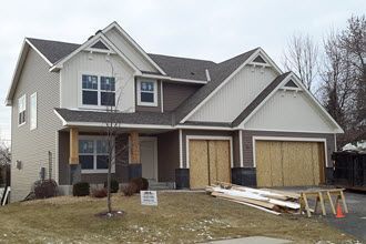 A two-story house under construction with tan siding, brown stone accents, and plywood covering the garage doors.