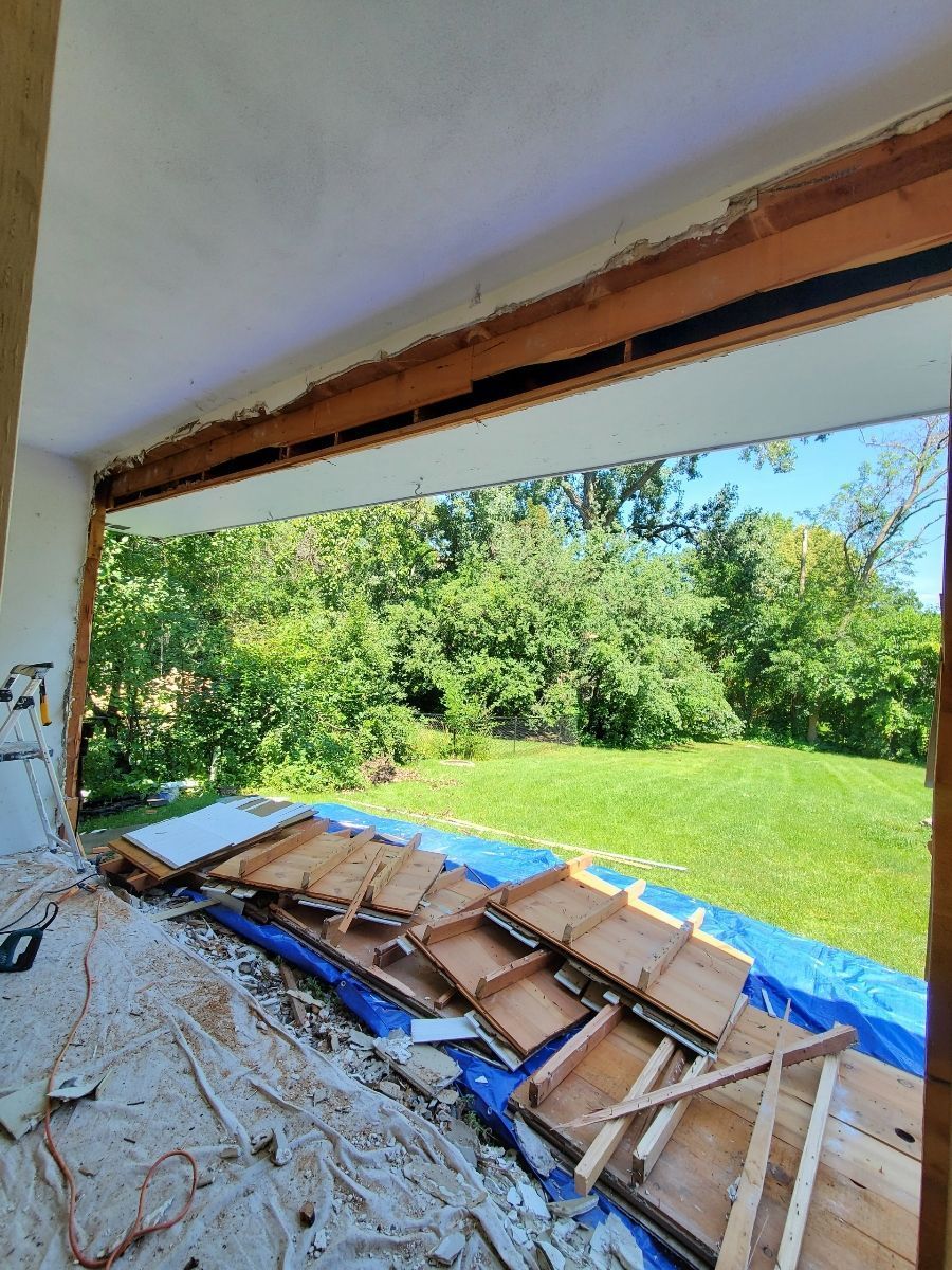 Interior view of a damaged wall opening to a green yard, with debris and wood framing on the floor