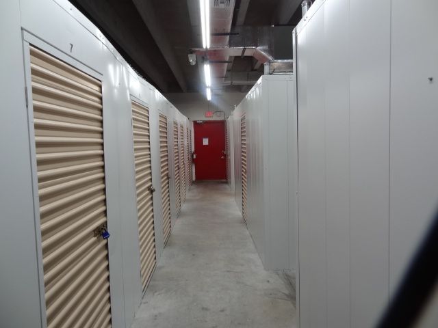 Interior of a storage facility; a hallway with rows of beige storage unit doors. Red door at end.