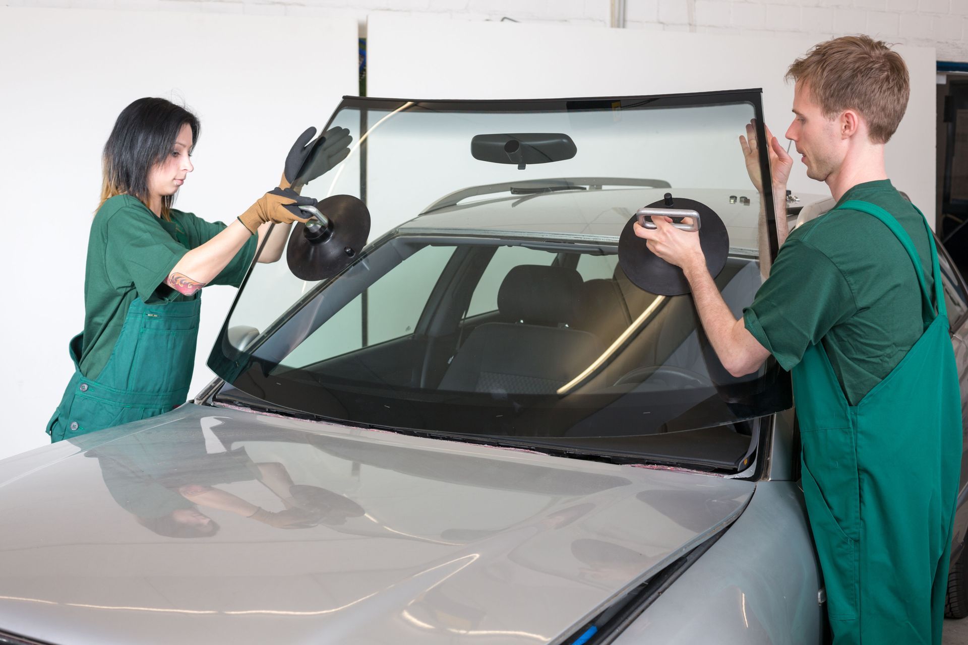 Two people installing a windshield on a silver car in a garage
