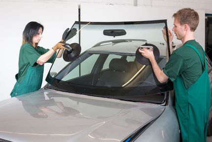 Two people installing a windshield on a silver car in a garage