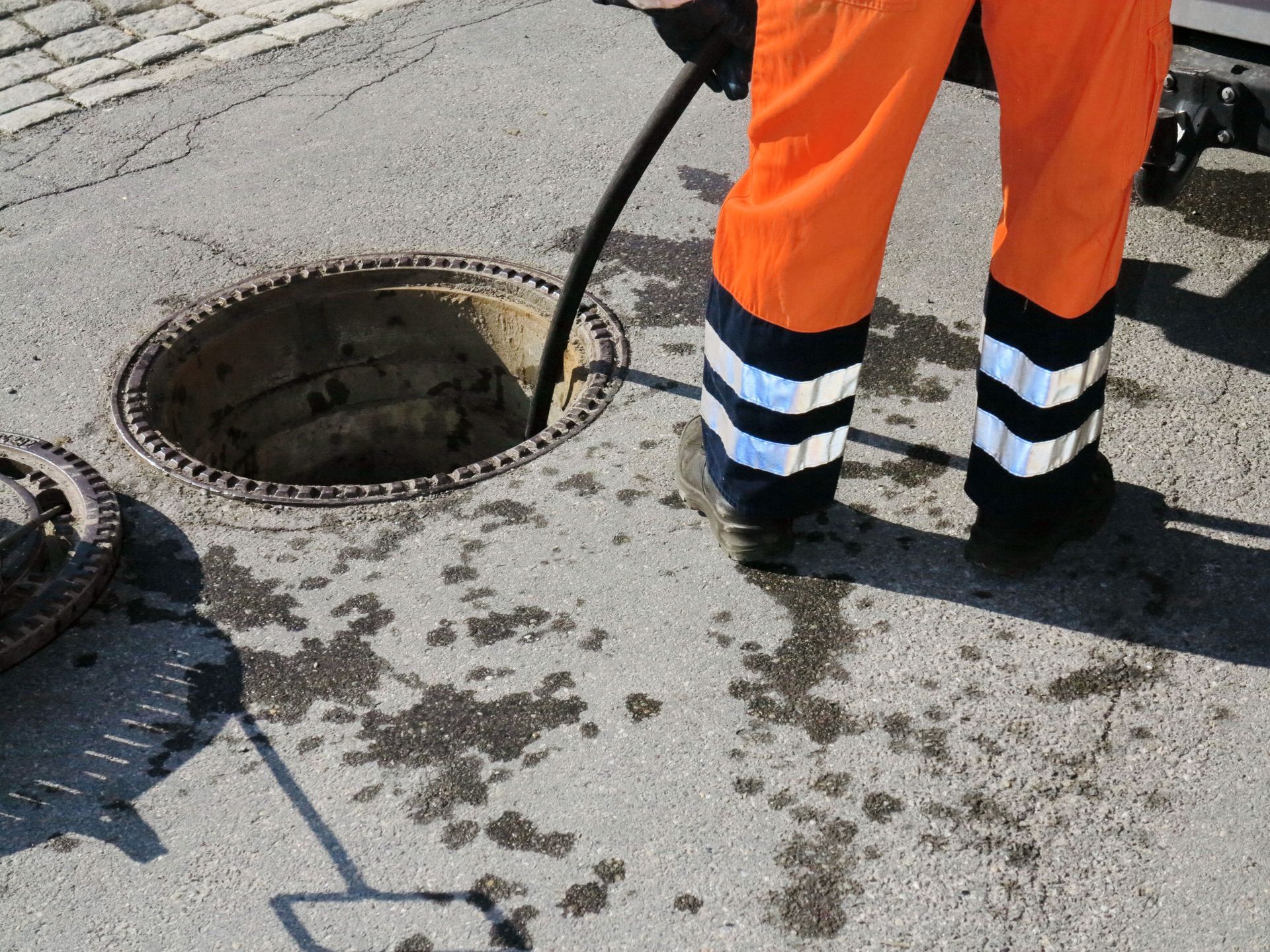Person in orange workwear, using a hose to work on a manhole cover in the street.
