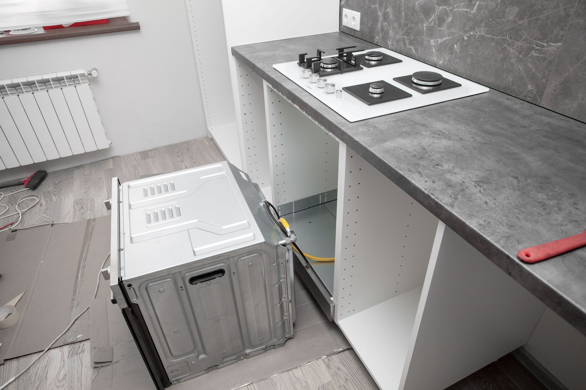 Kitchen renovation in progress: oven being installed in white cabinetry below a gray countertop with a stovetop.