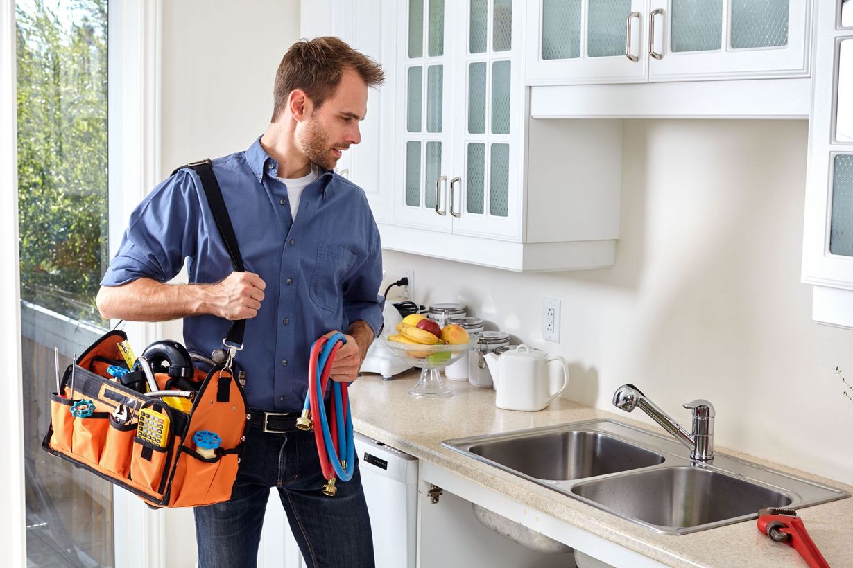 Plumber with tool bag examines kitchen sink.