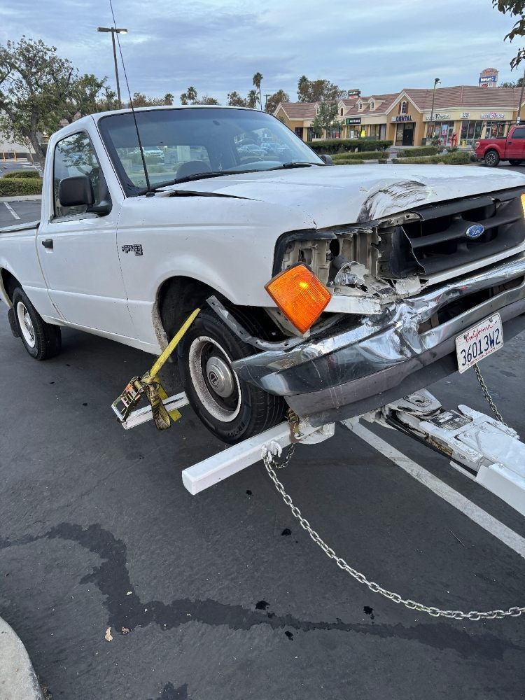 White pickup truck with severe front-end damage, missing grille, and a bent tow bar in a parking lot