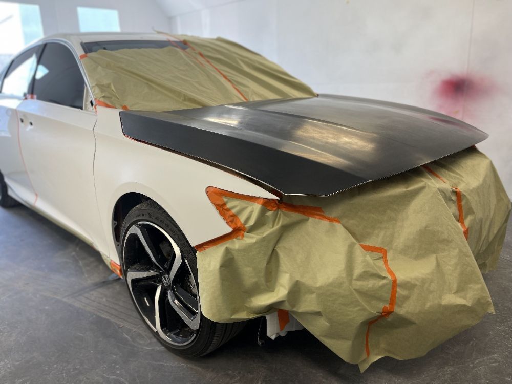 White sedan with black hood and wheels, masked with paper and tape in a paint booth