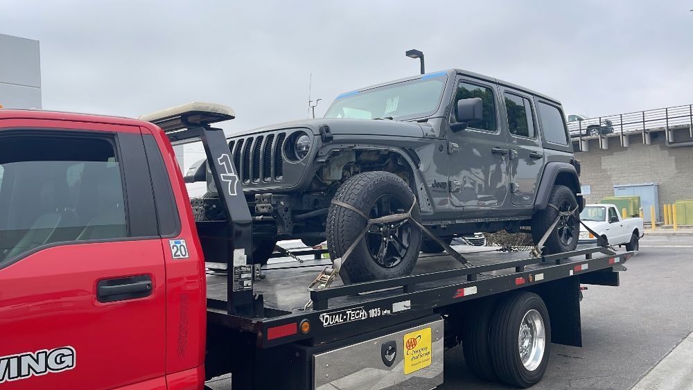 Black Jeep Wrangler on a flatbed tow truck beside a red truck on a highway.