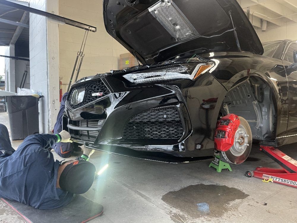 Black sports car on a lift in a garage, hood open with mechanic working underneath the front bumper