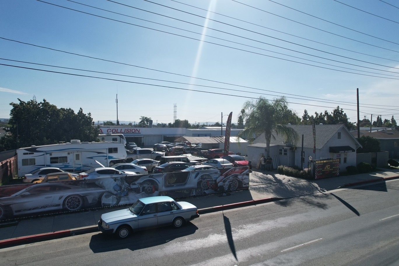 Sunlit parking lot crowded with cars beside low buildings and palm trees, viewed from a roadside.