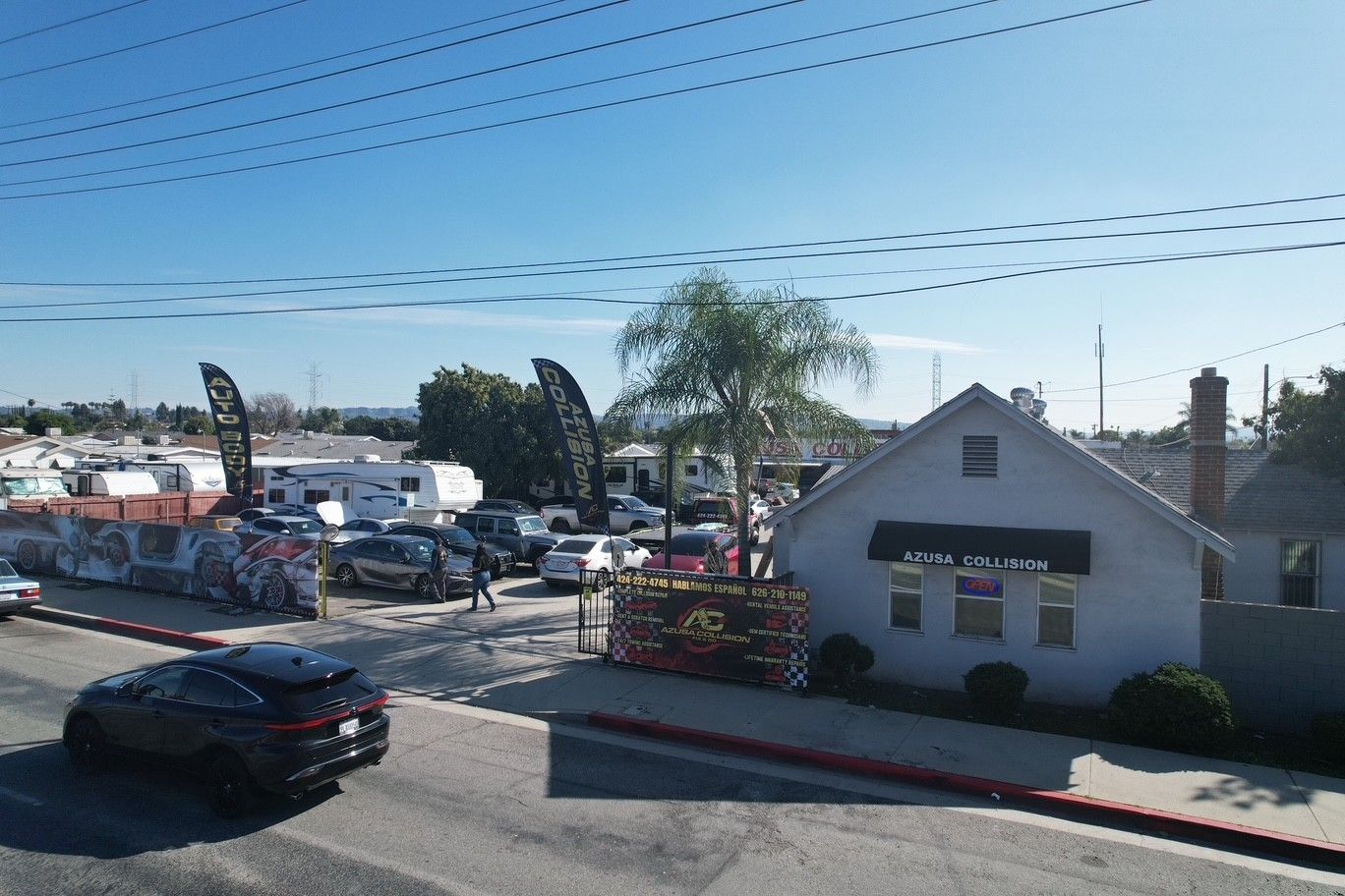 Car lot beside a white building and palm tree under a clear sky, with parked cars and utility wires.