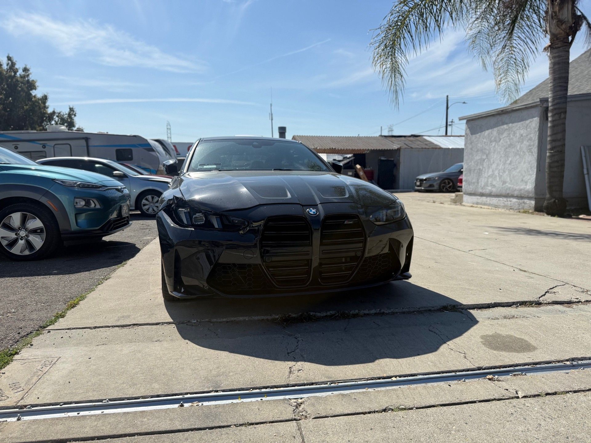 Black sports sedan parked in a sunny lot between other cars, with a palm tree and building nearby