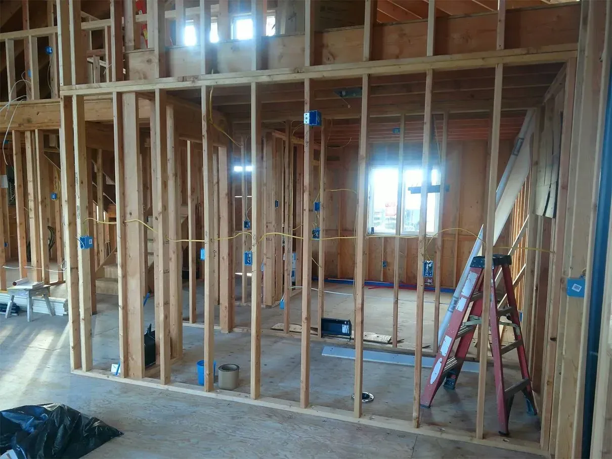 Interior view of a building under construction, showing wooden framing, electrical boxes, and a ladder.