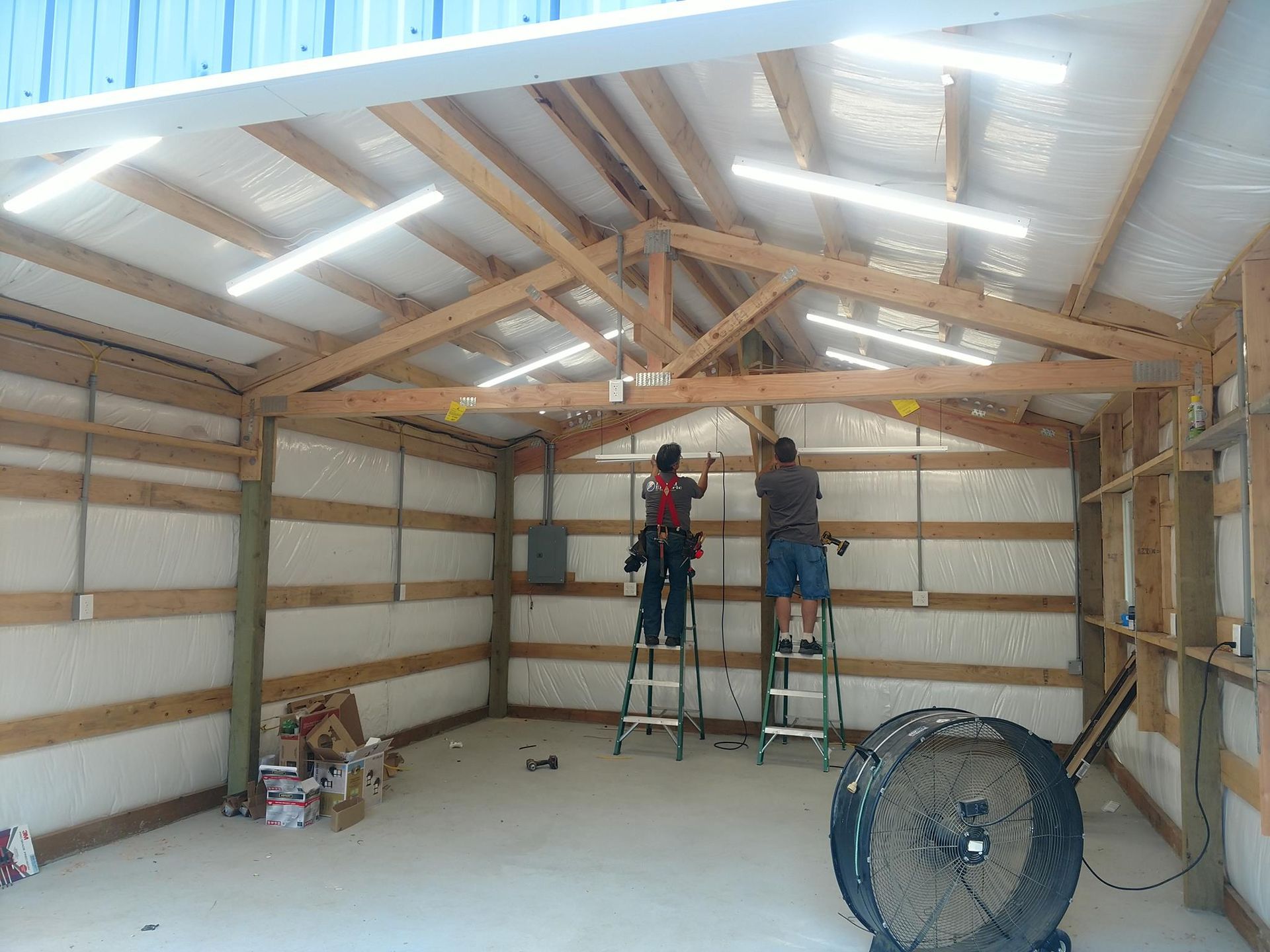Two people on ladders installing lights in a wood-framed garage with insulation and a concrete floor.