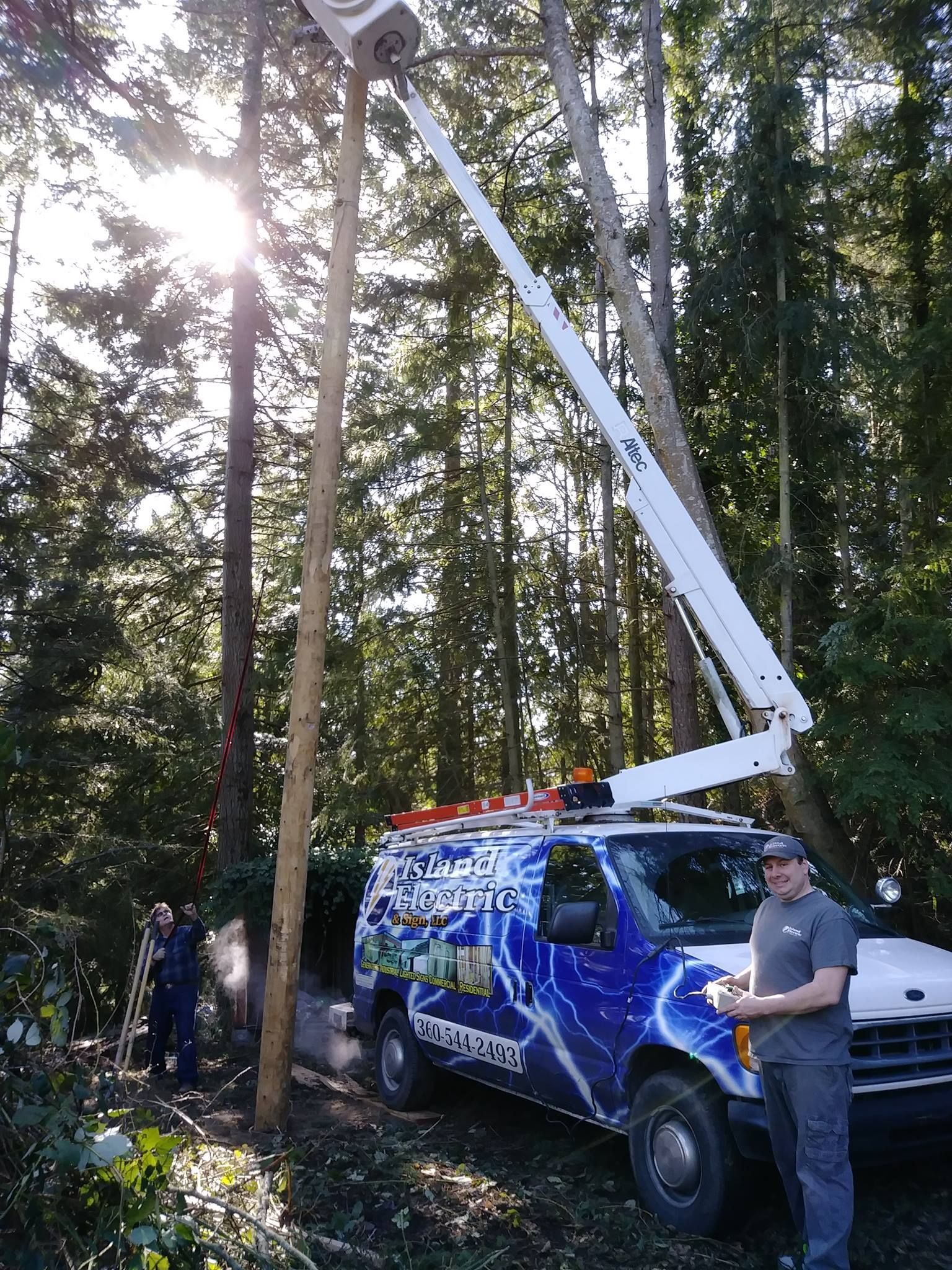 Tree service truck with lift trimming a tall tree in a wooded area; person operating controls.
