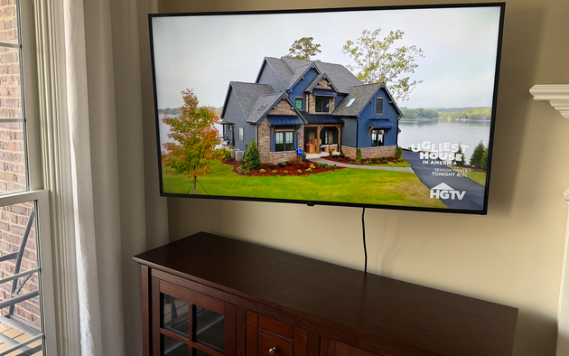 A large flat-screen TV mounted on a wall above a dark wooden cabinet, displaying an HGTV home exterior program.