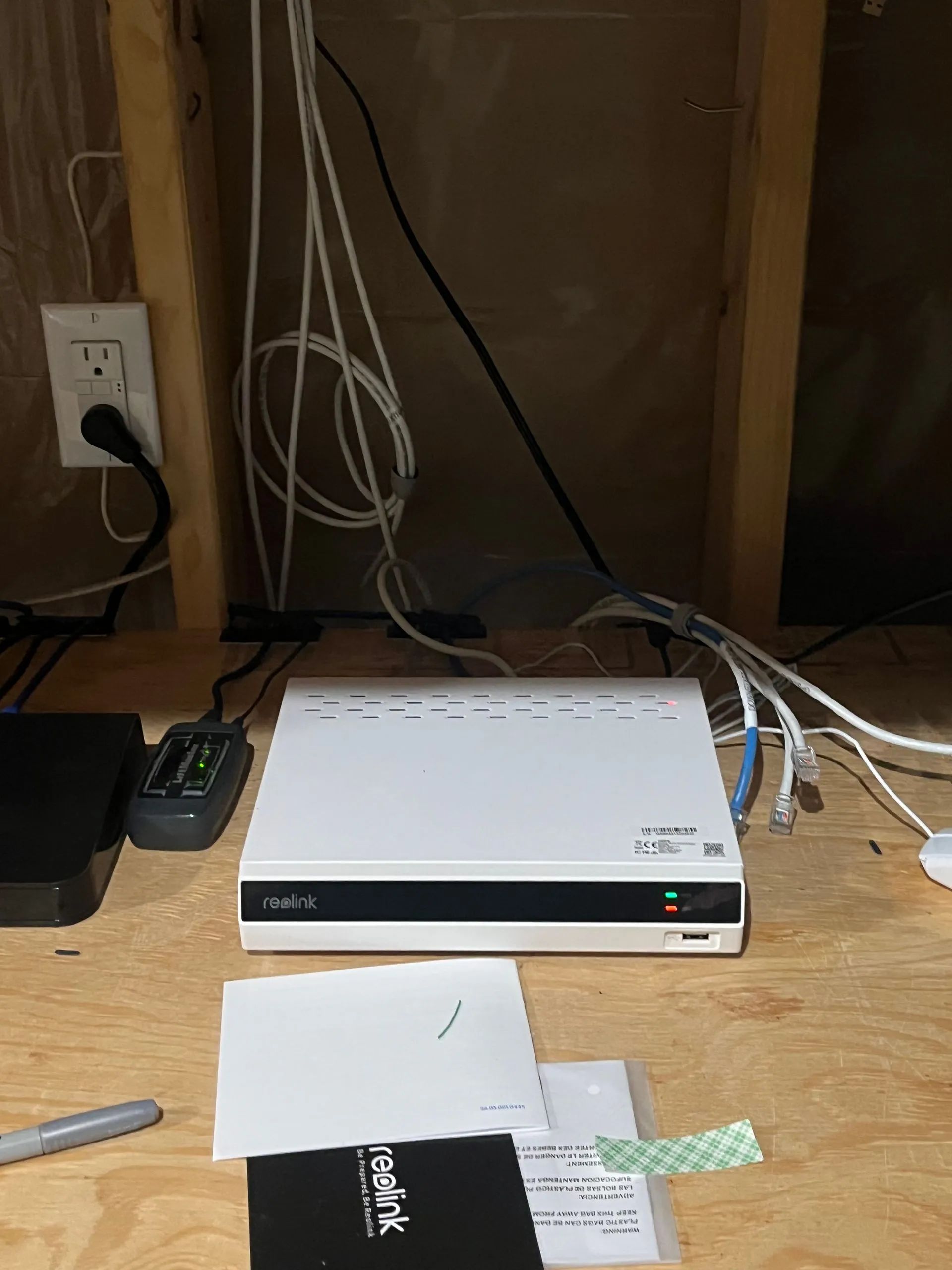 A Reolink NVR device sits on a wooden surface with connected ethernet cables and power cords, surrounded by manuals.
