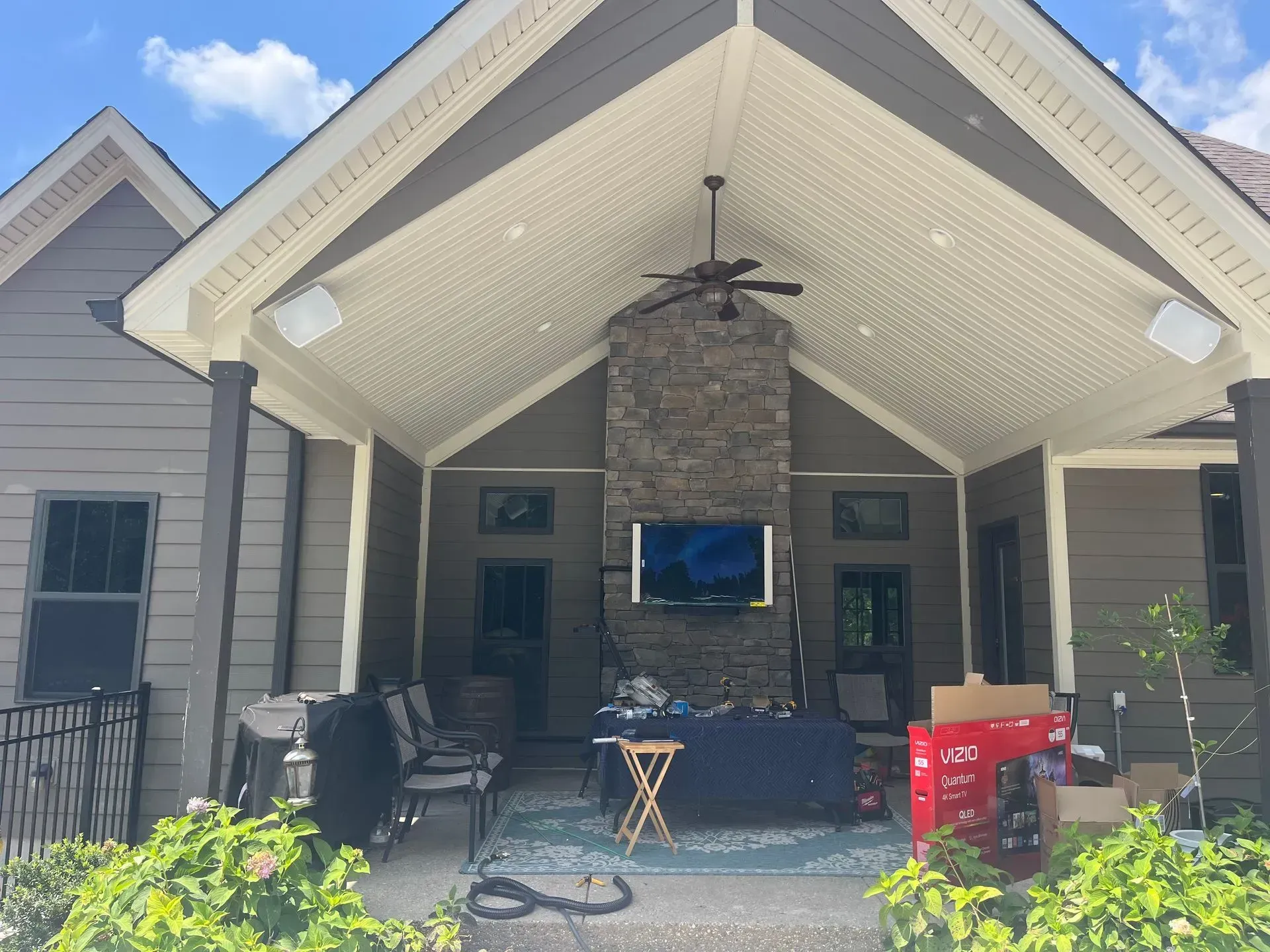 A covered backyard patio with a stone fireplace, mounted television, ceiling fan, outdoor furniture, and a box of goods.