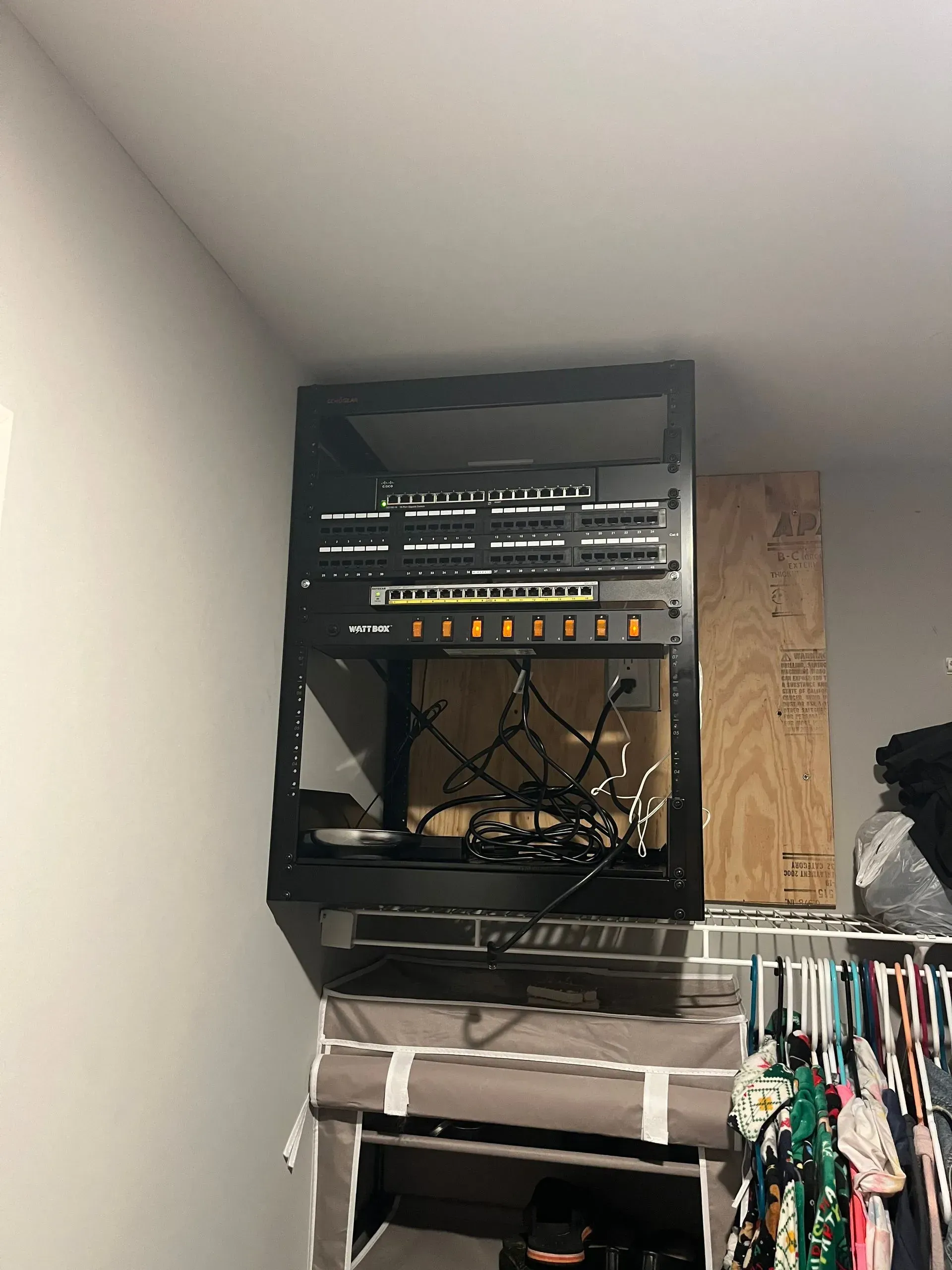 A black wall-mounted server rack holding networking equipment, installed in a closet above a clothing rack.