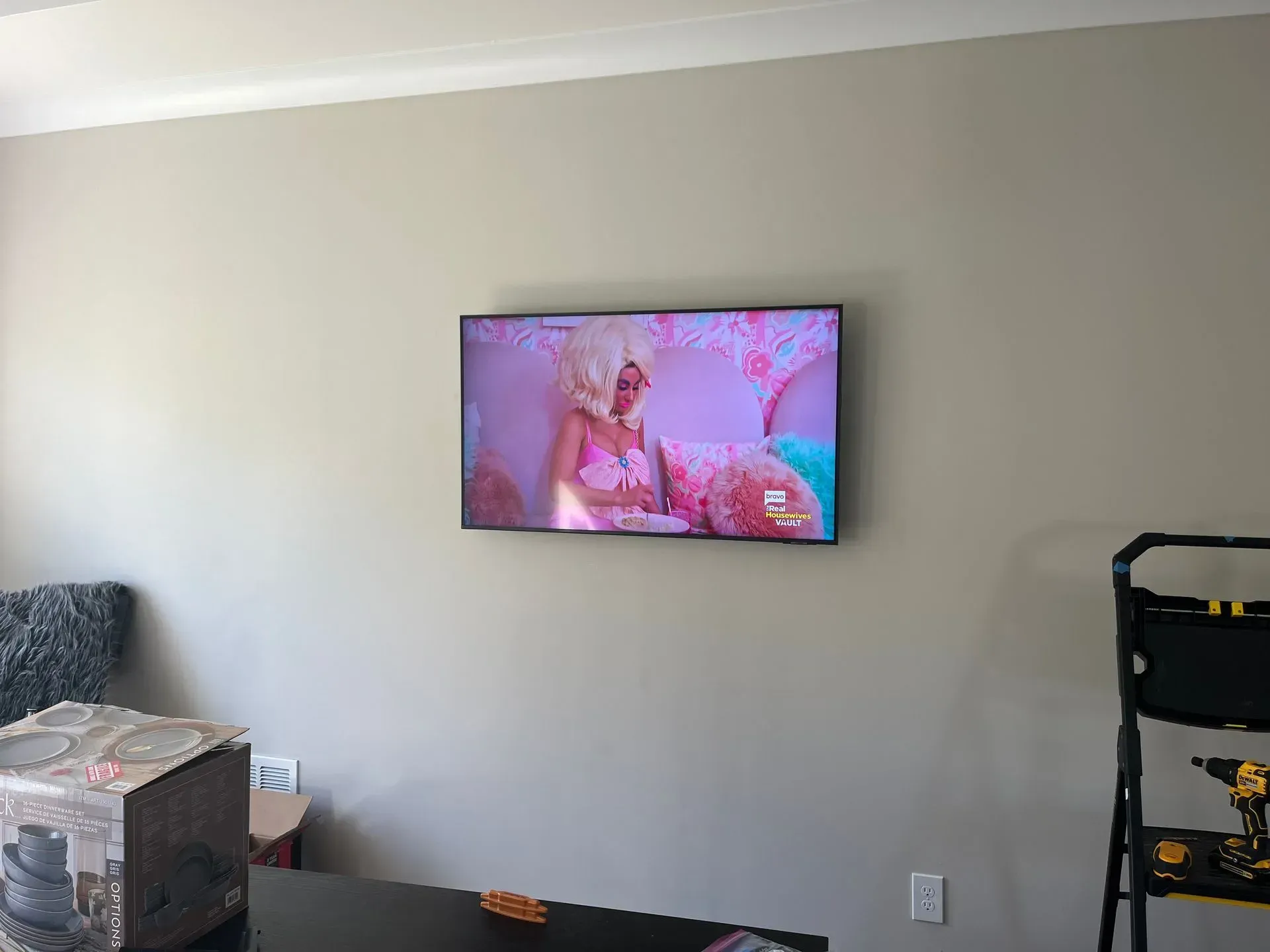 A television mounted on a beige wall displays a scene with pink and white décor; a ladder and a box are in the room.