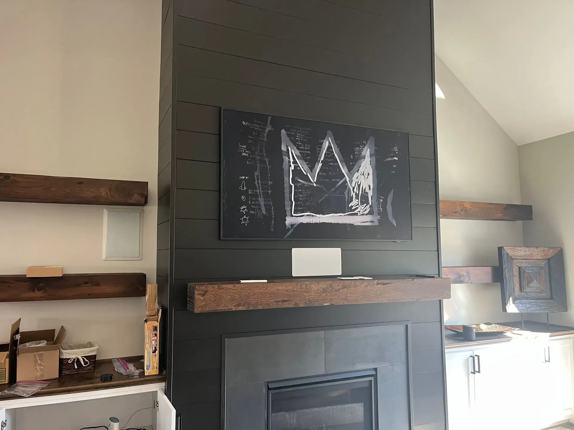 A black-painted fireplace with horizontal shiplap, a rustic wood mantel, and a crown-motif artwork hanging above it.