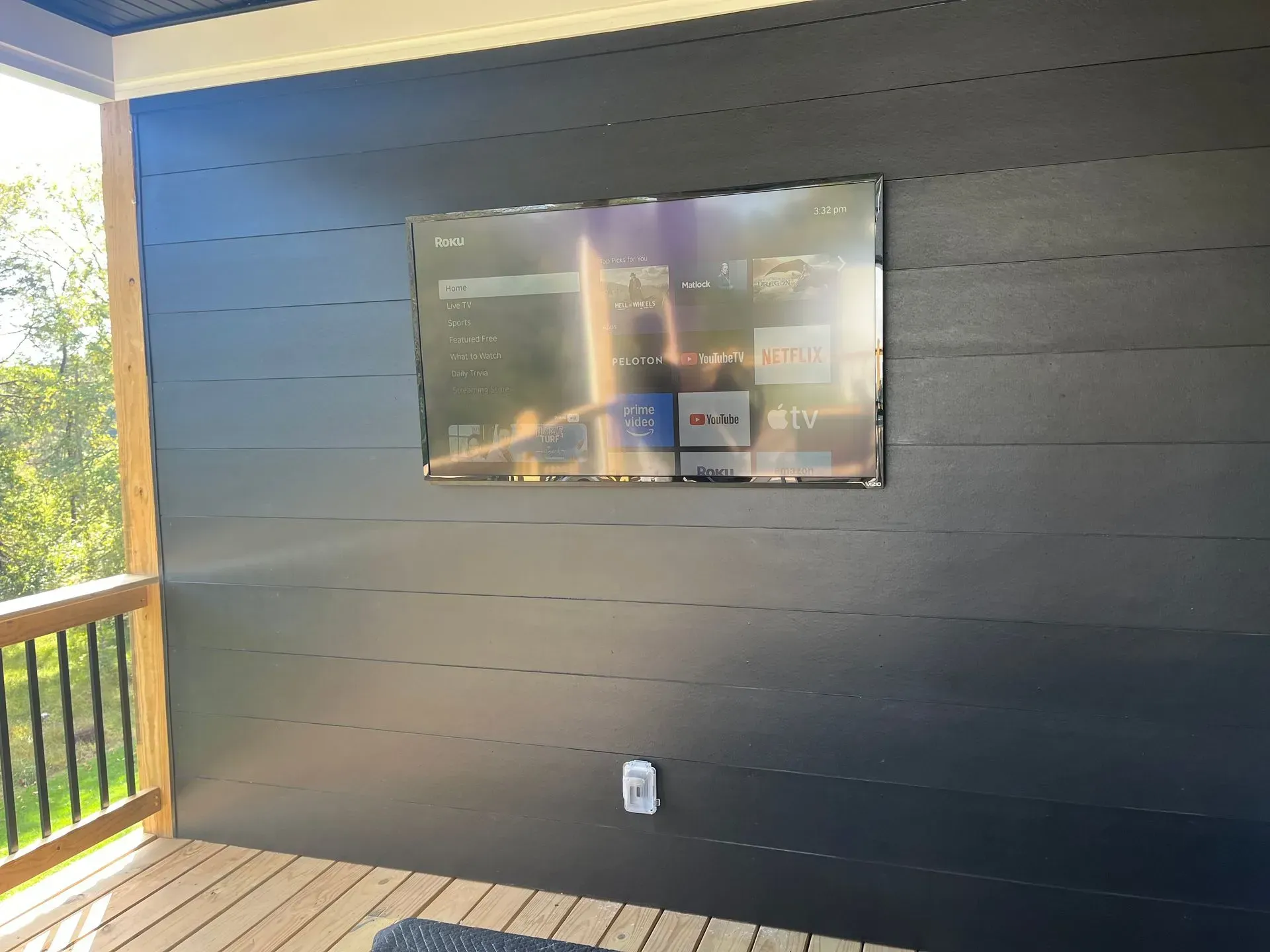 A television mounted on a dark horizontal paneled wall on an outdoor wooden deck with a railing.
