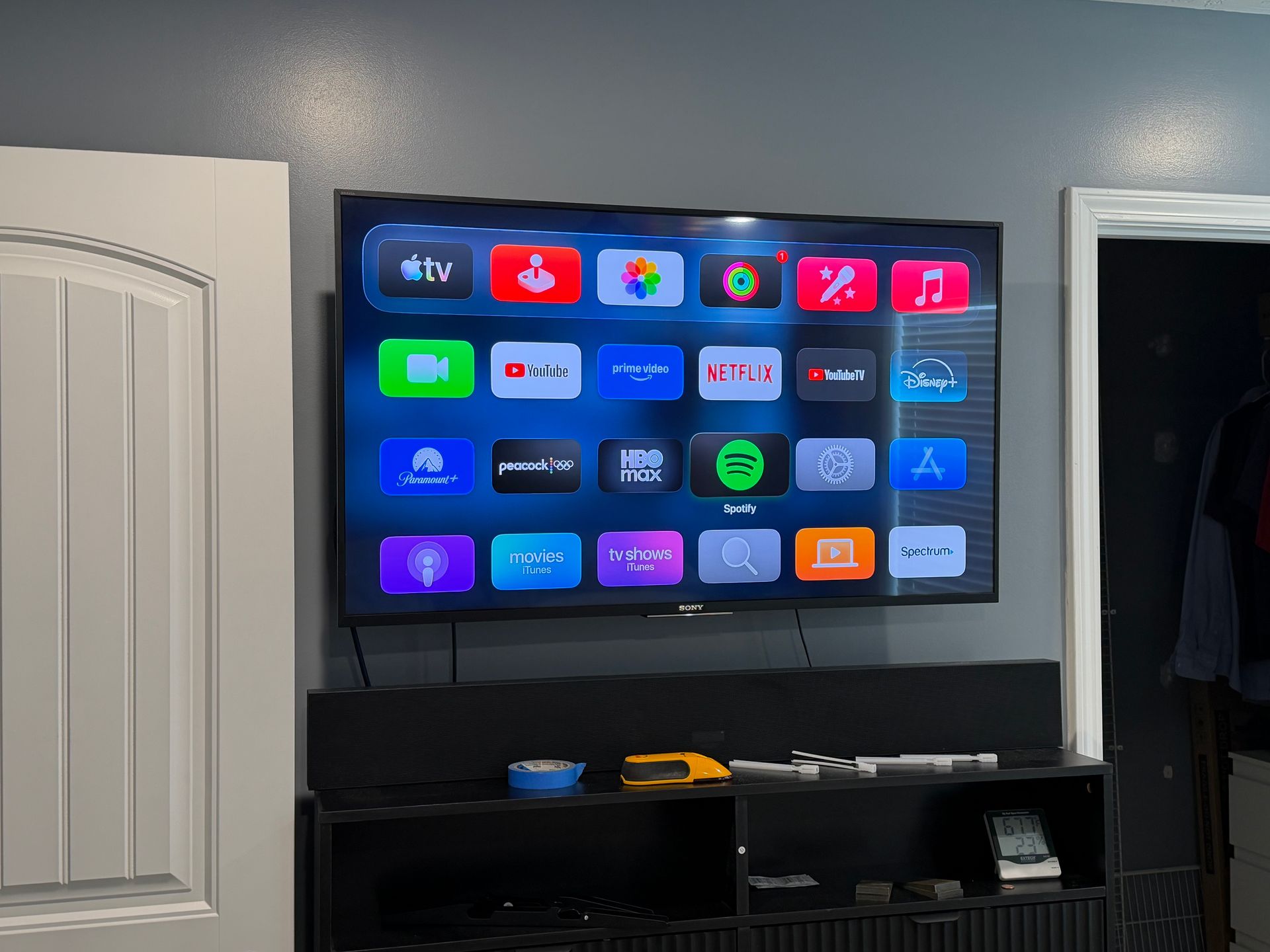 A flat-screen TV mounted on a gray wall above a black console, displaying an Apple TV home screen with app icons.