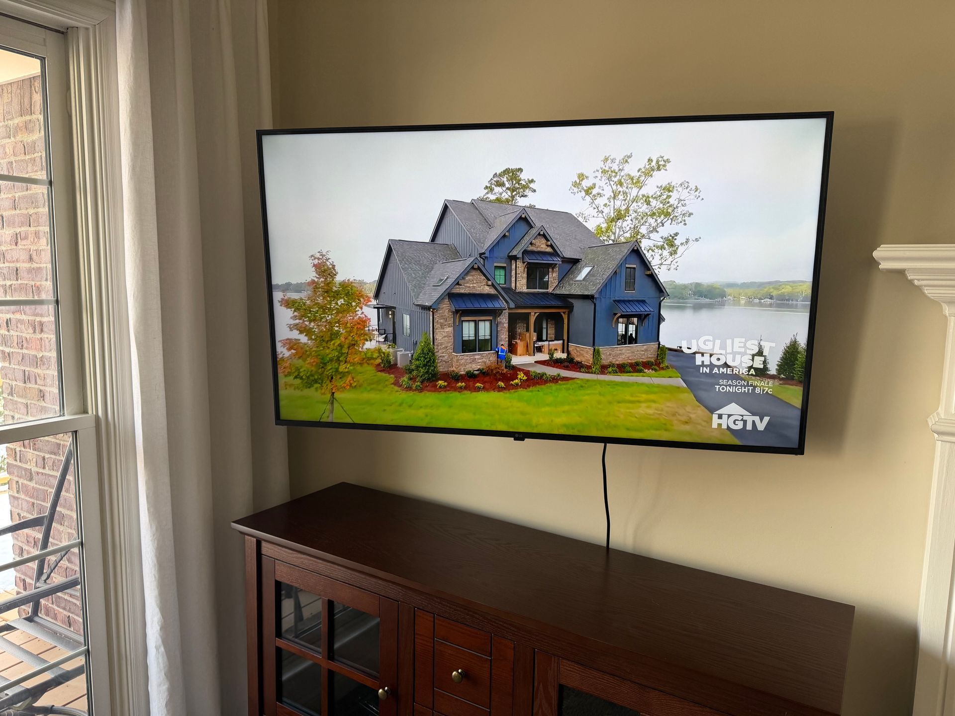 A flat-screen television mounted on a wall above a dark wooden cabinet, displaying a house image from HGTV.