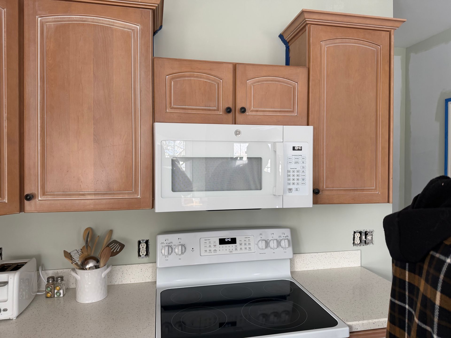 A white microwave mounted between two light-wood cabinets above a white electric stove in a kitchen.