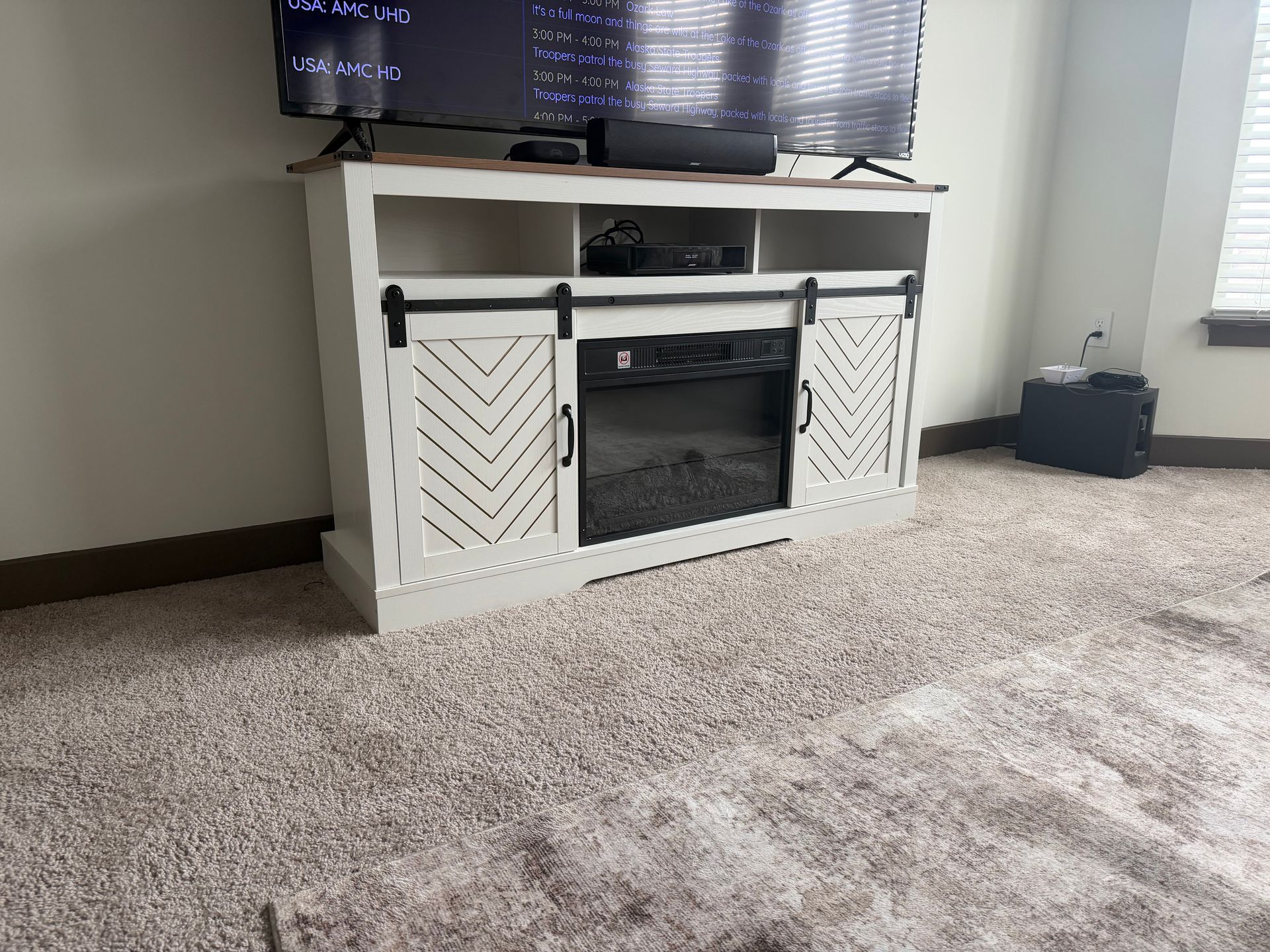 A white TV stand with sliding barn doors and an electric fireplace, situated on a carpeted floor below a wall-mounted TV.