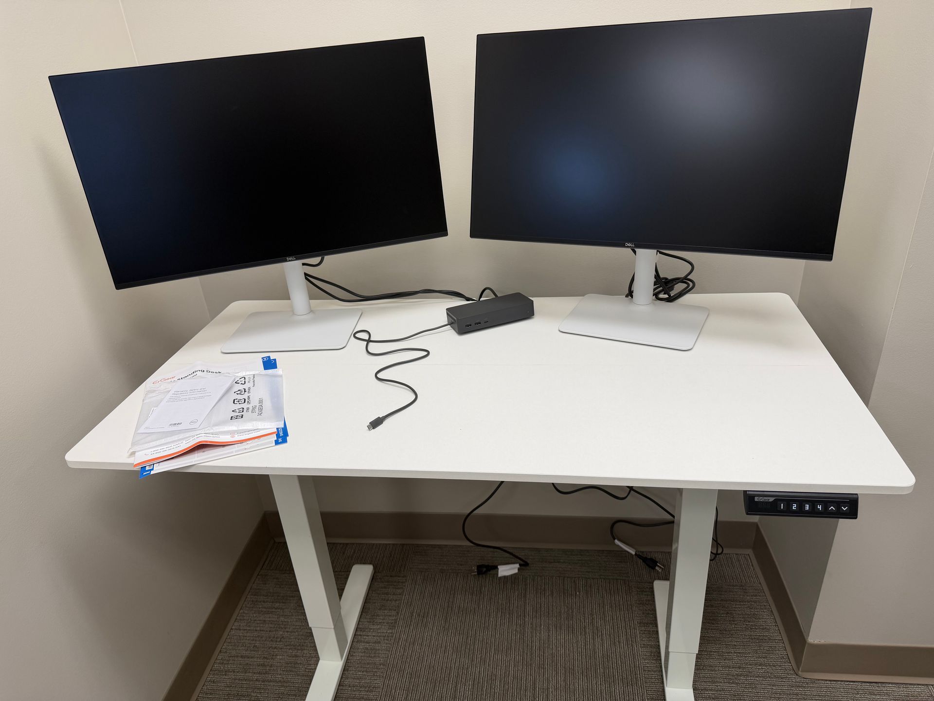 A white standing desk with two black monitors, a power brick, and a small stack of papers against a light-colored wall.