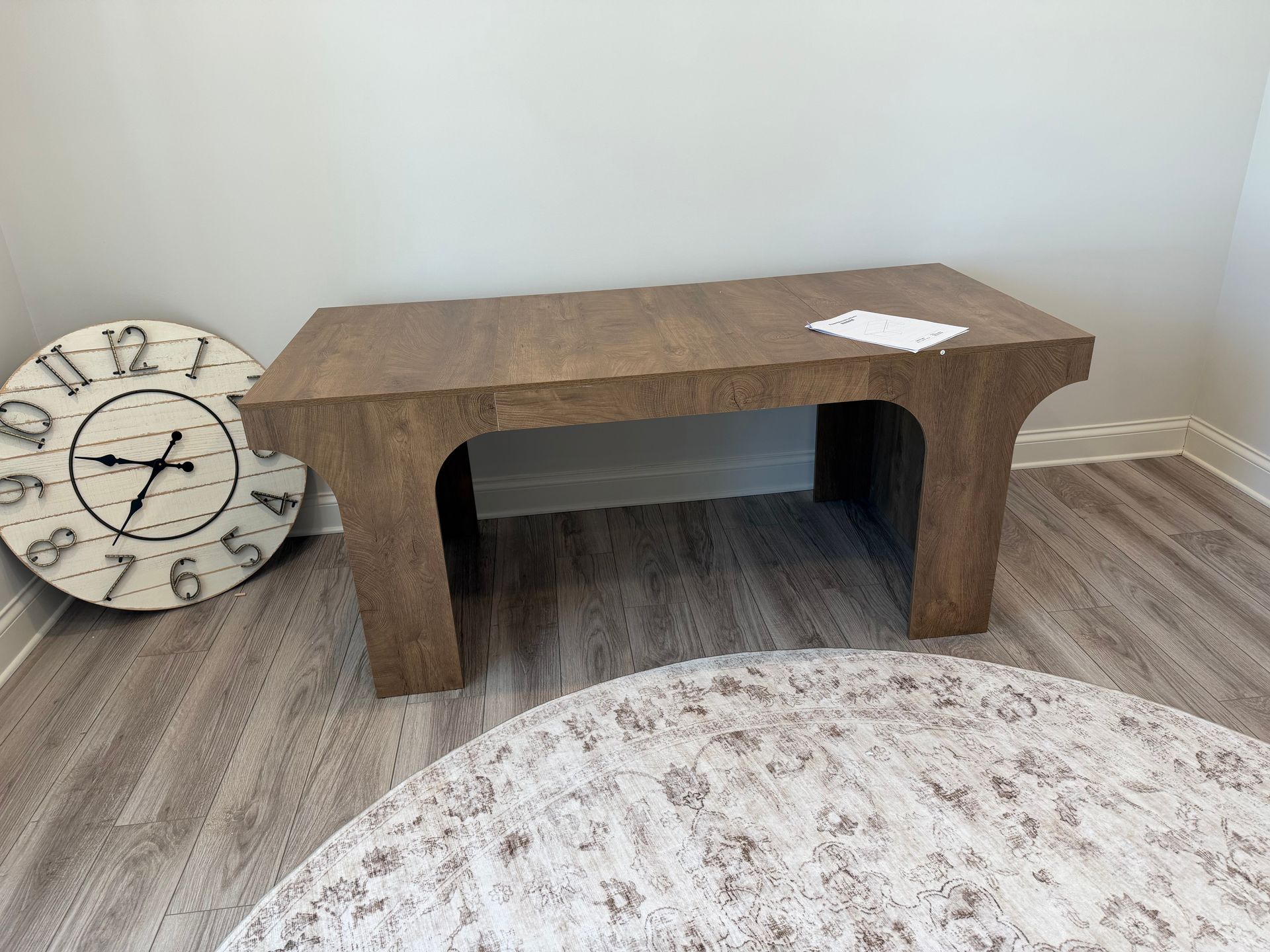 A rustic wooden bench with arched legs stands in a room with a large wall clock and a patterned rug on the floor.