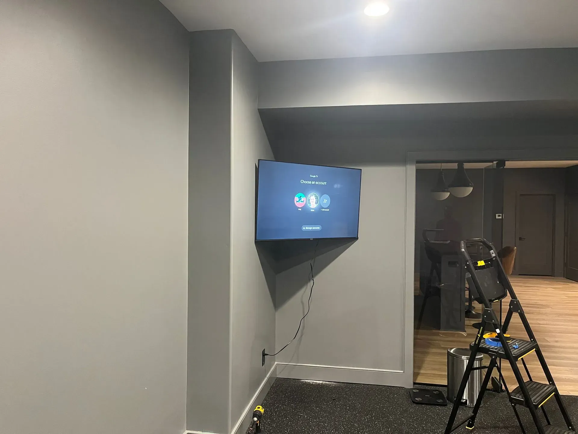 A wall-mounted TV displays a menu in a grey room with a step ladder nearby.