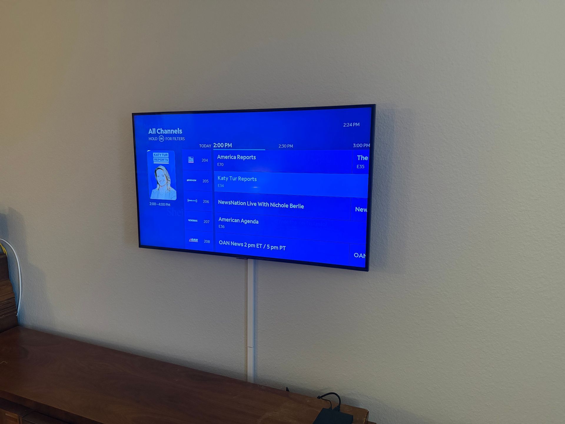 A flat-screen TV mounted on a beige wall displays a blue menu, with a cable cover running down to a wooden surface below.