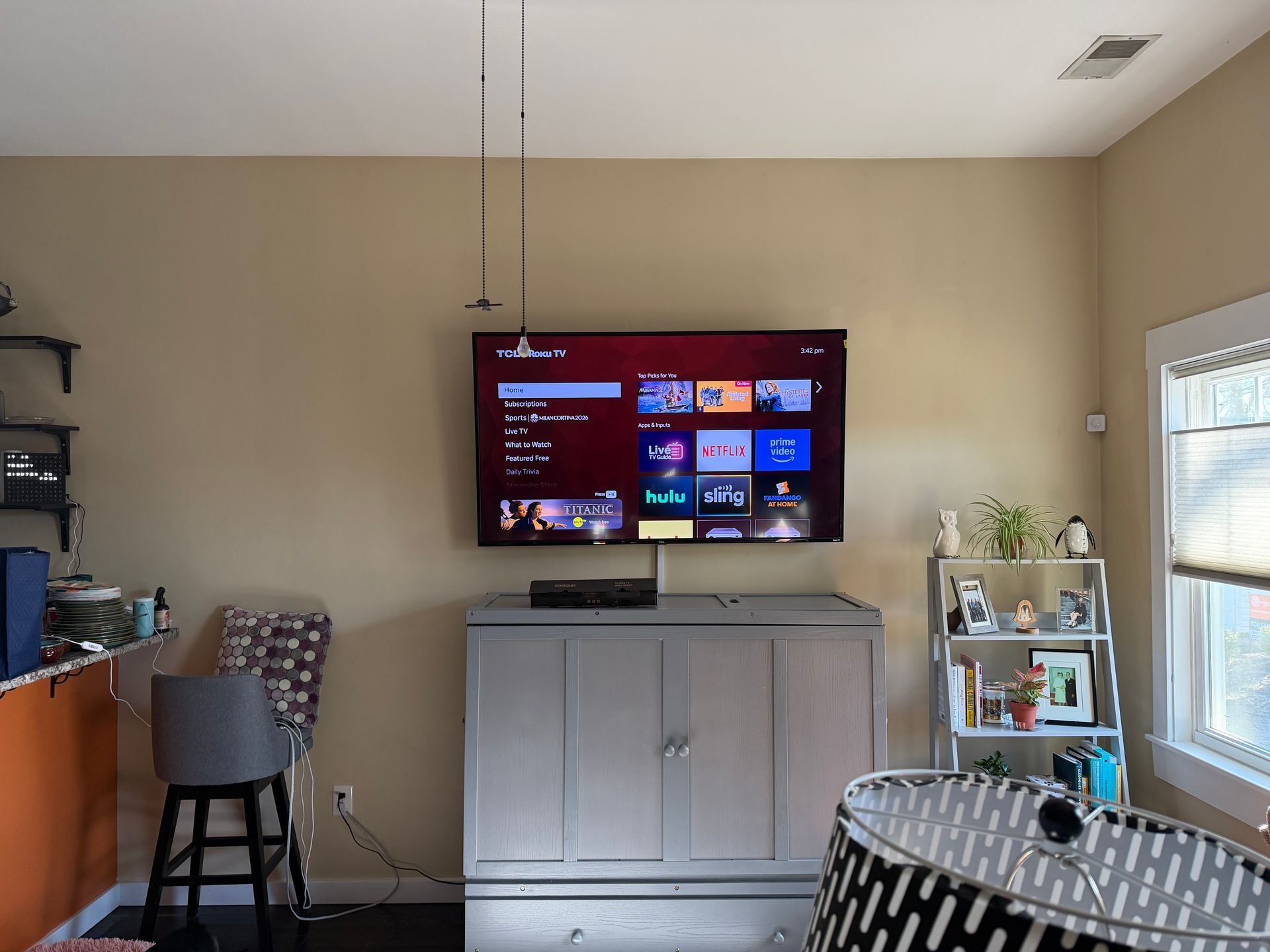 A living room featuring a mounted television above a gray cabinet, with a stool to the left and a shelf unit on the right.