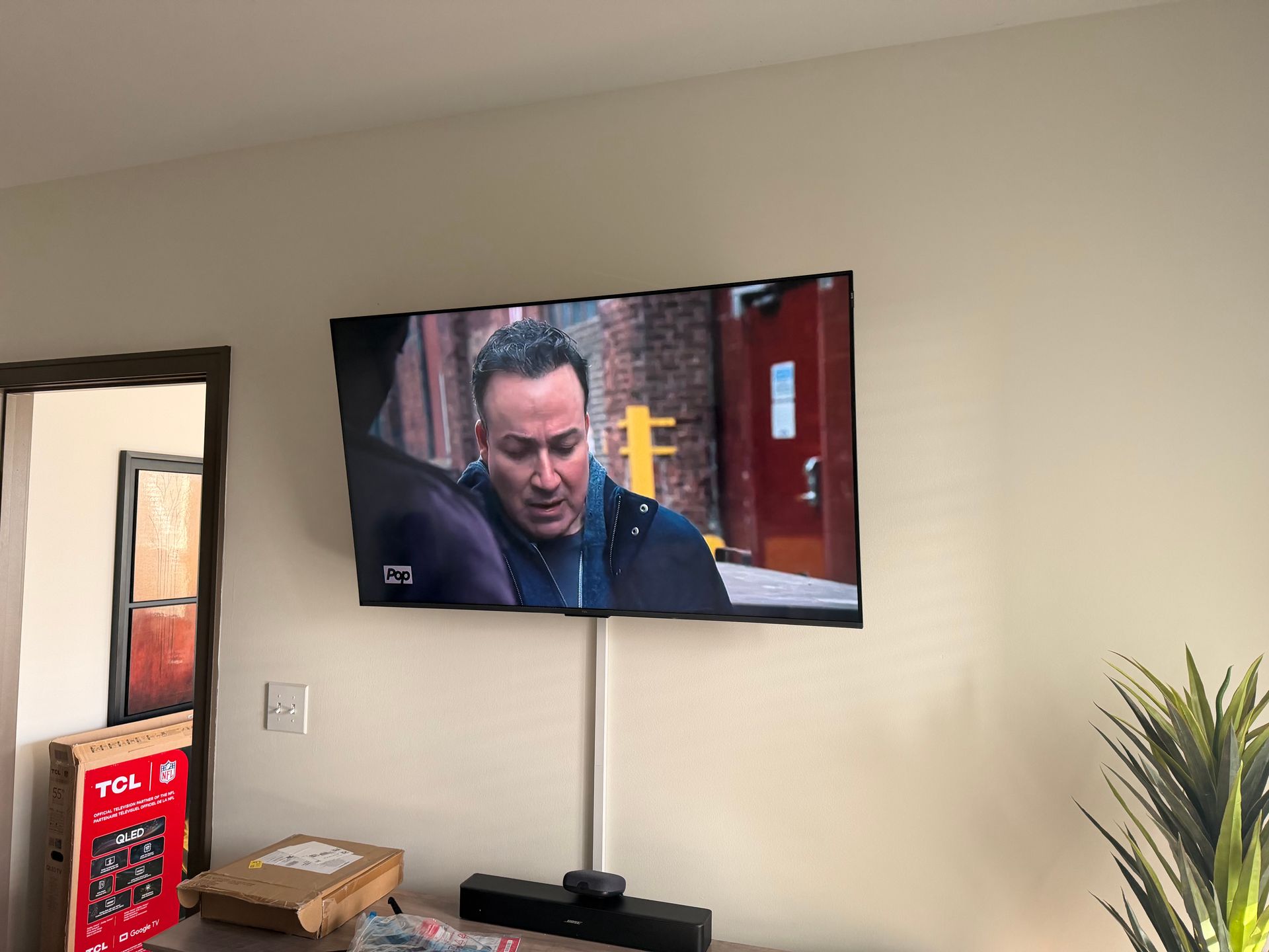 A flat-screen TV mounted on a light-colored wall with a cable cover, showing a man on screen, above a soundbar.