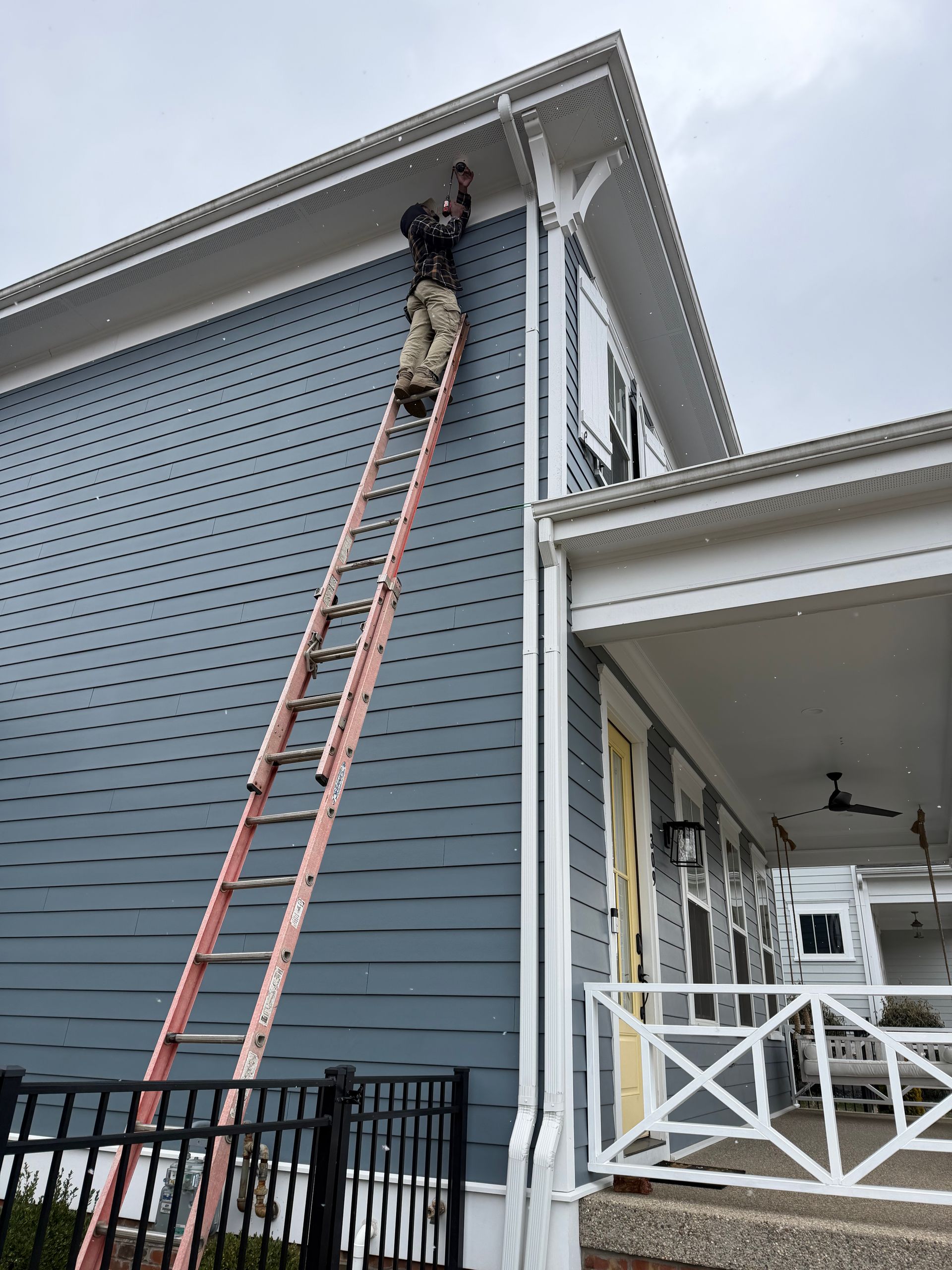 A person on a tall ladder reaches toward the roofline of a blue-sided house with a white-railed porch.