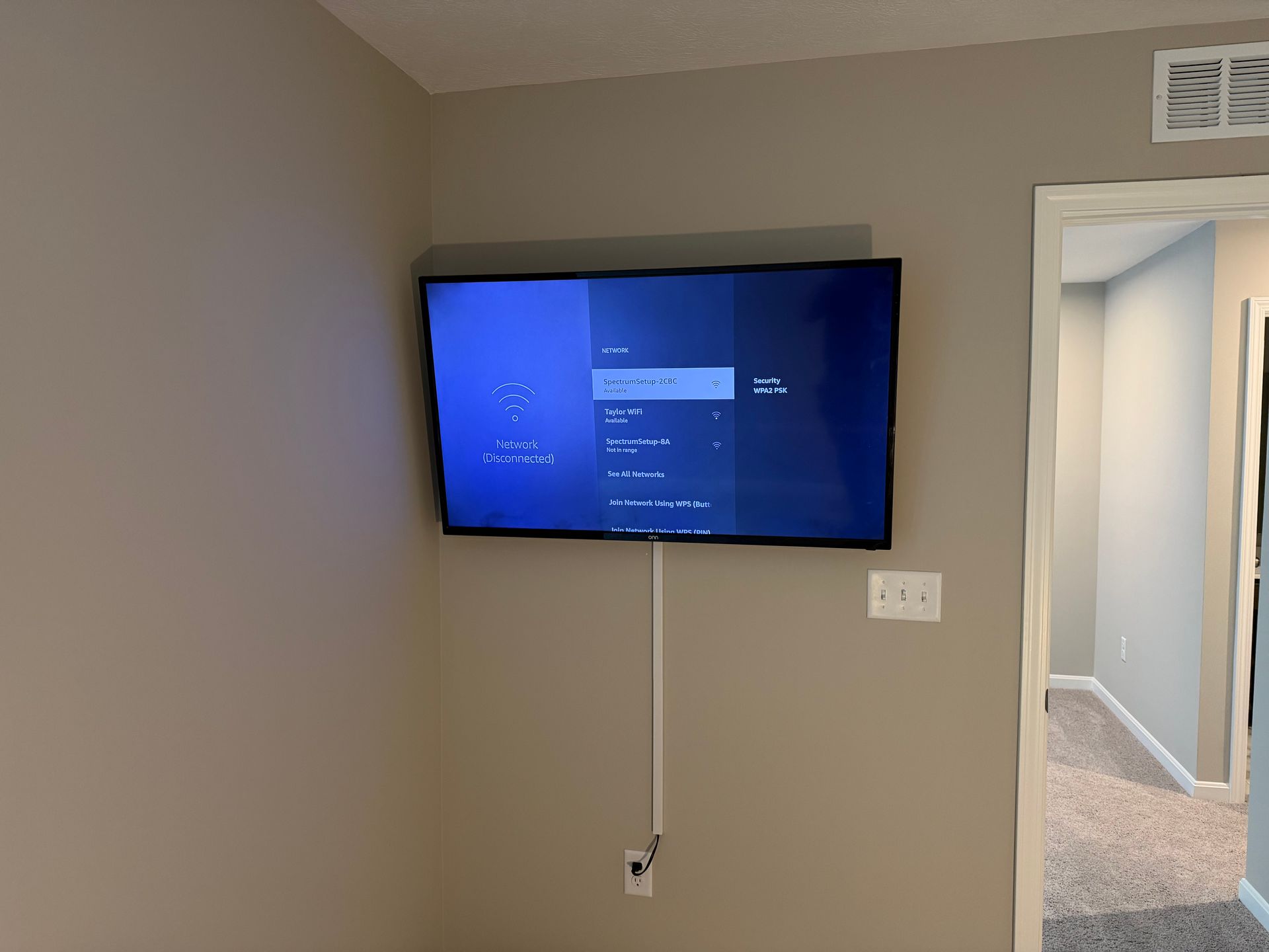 A television mounted on a beige wall displays a blue menu screen with a white cable cover leading to a power outlet below.