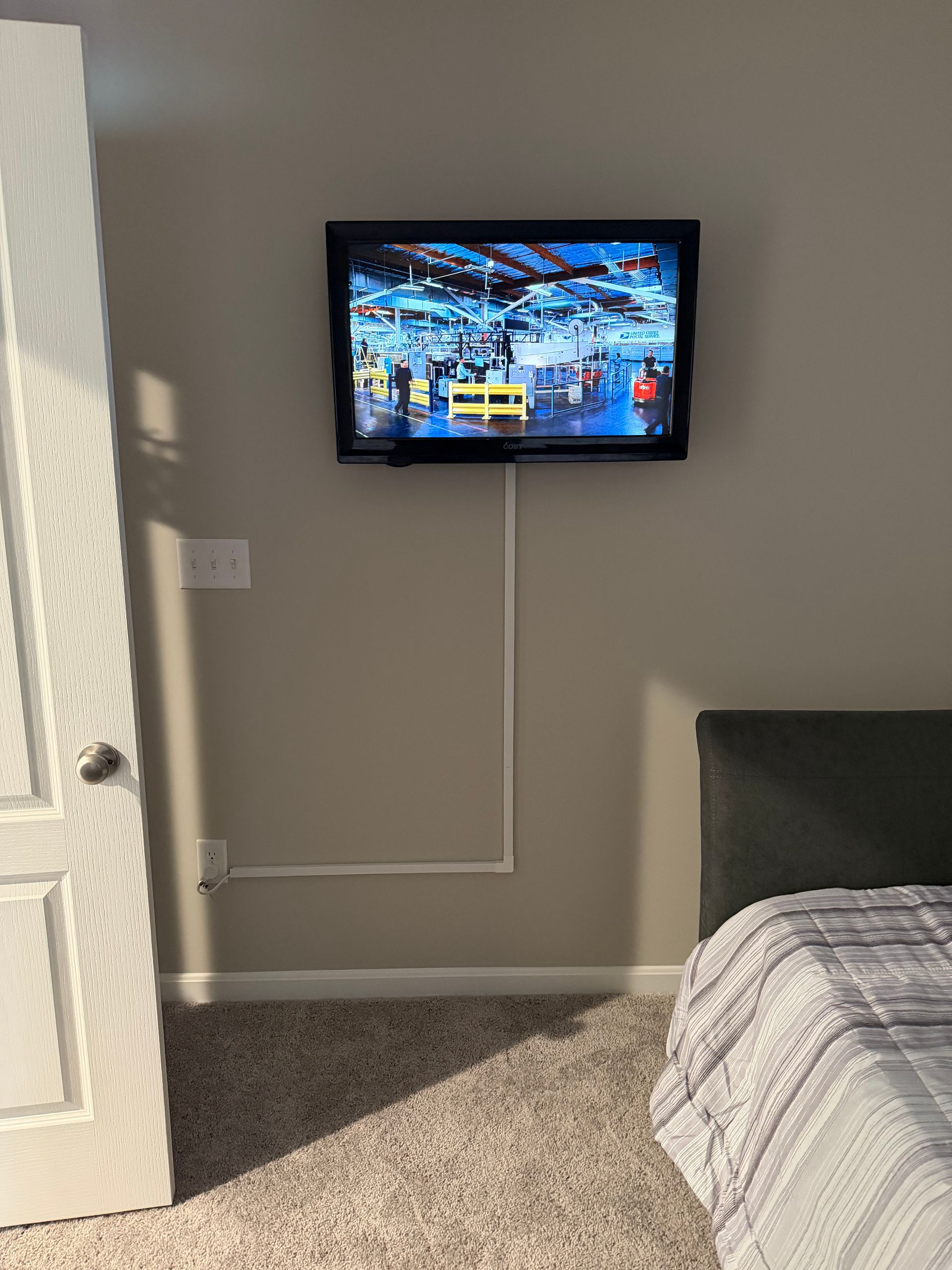 A flat-screen TV mounted on a beige wall above a bedroom carpet, with a white cord cover running down to an outlet.