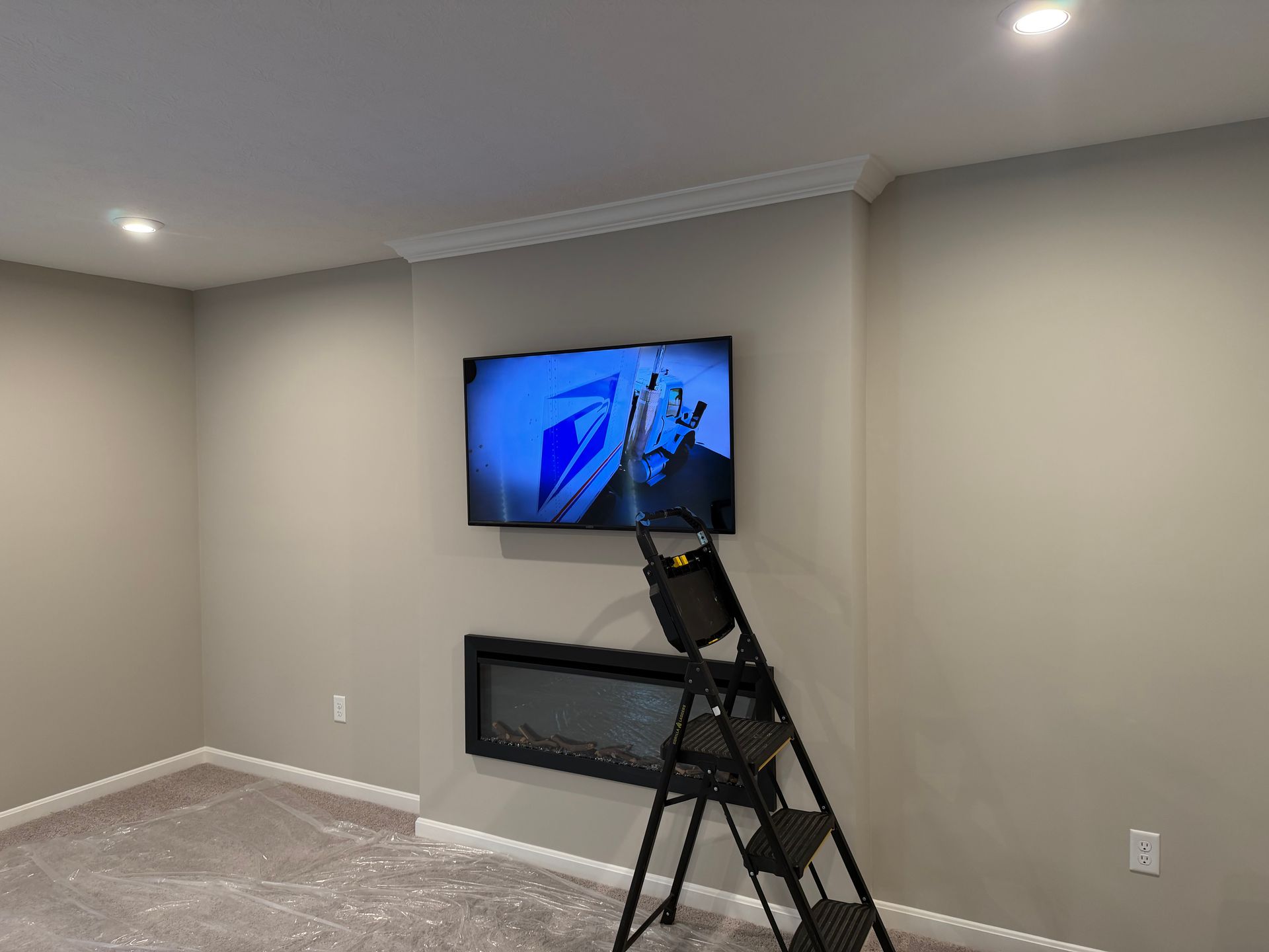 A black stepladder stands in front of a neutral-toned wall with a mounted flat-screen television and a linear fireplace.