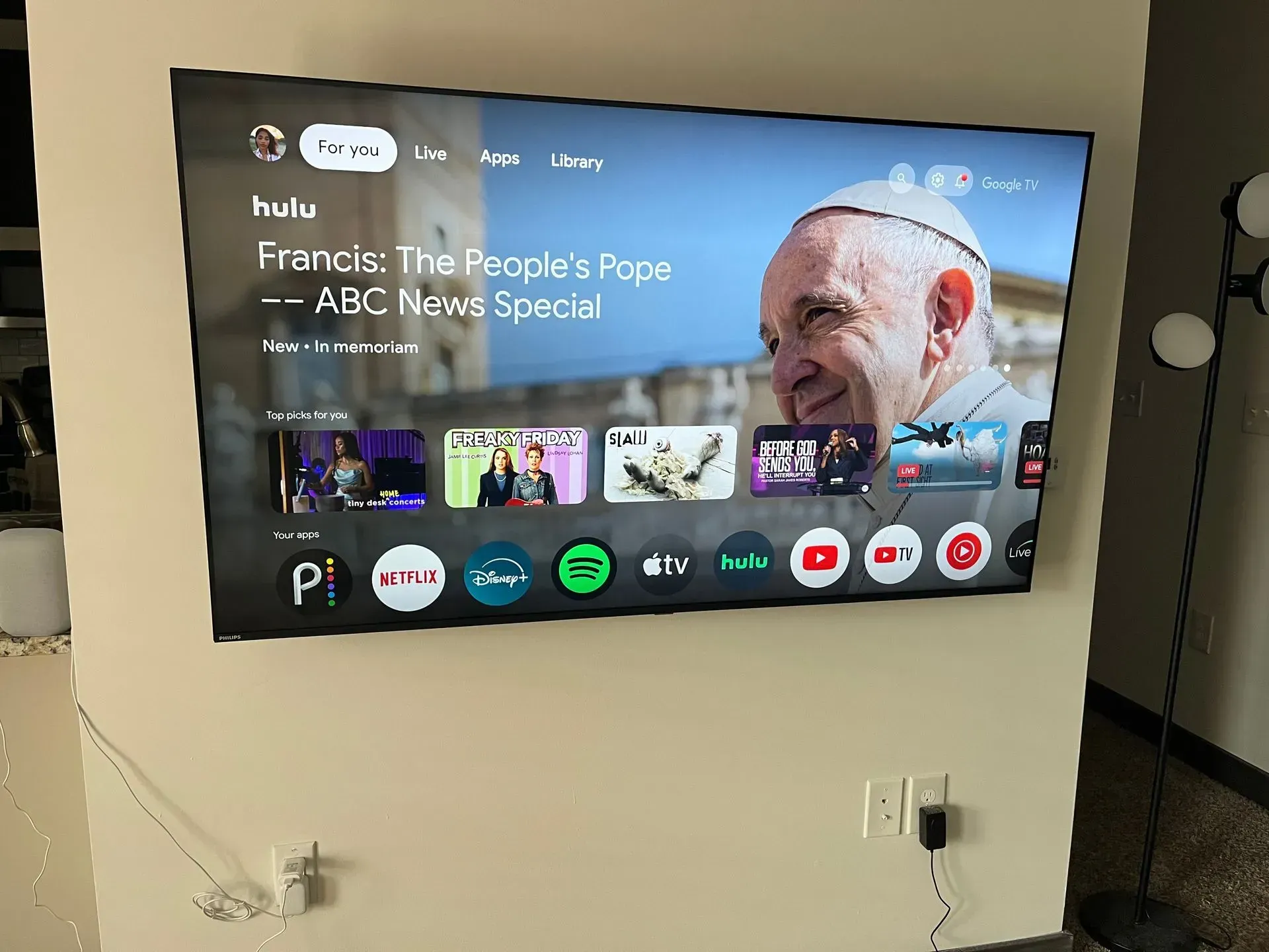 A wall-mounted TV showing a Google TV interface displaying an ABC News special about Pope Francis, with app icons below.