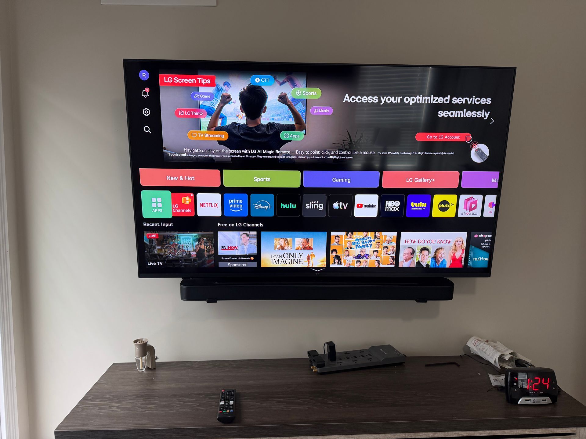 A wall-mounted TV showing a smart interface with various app icons, placed above a wooden table with a remote and clock.
