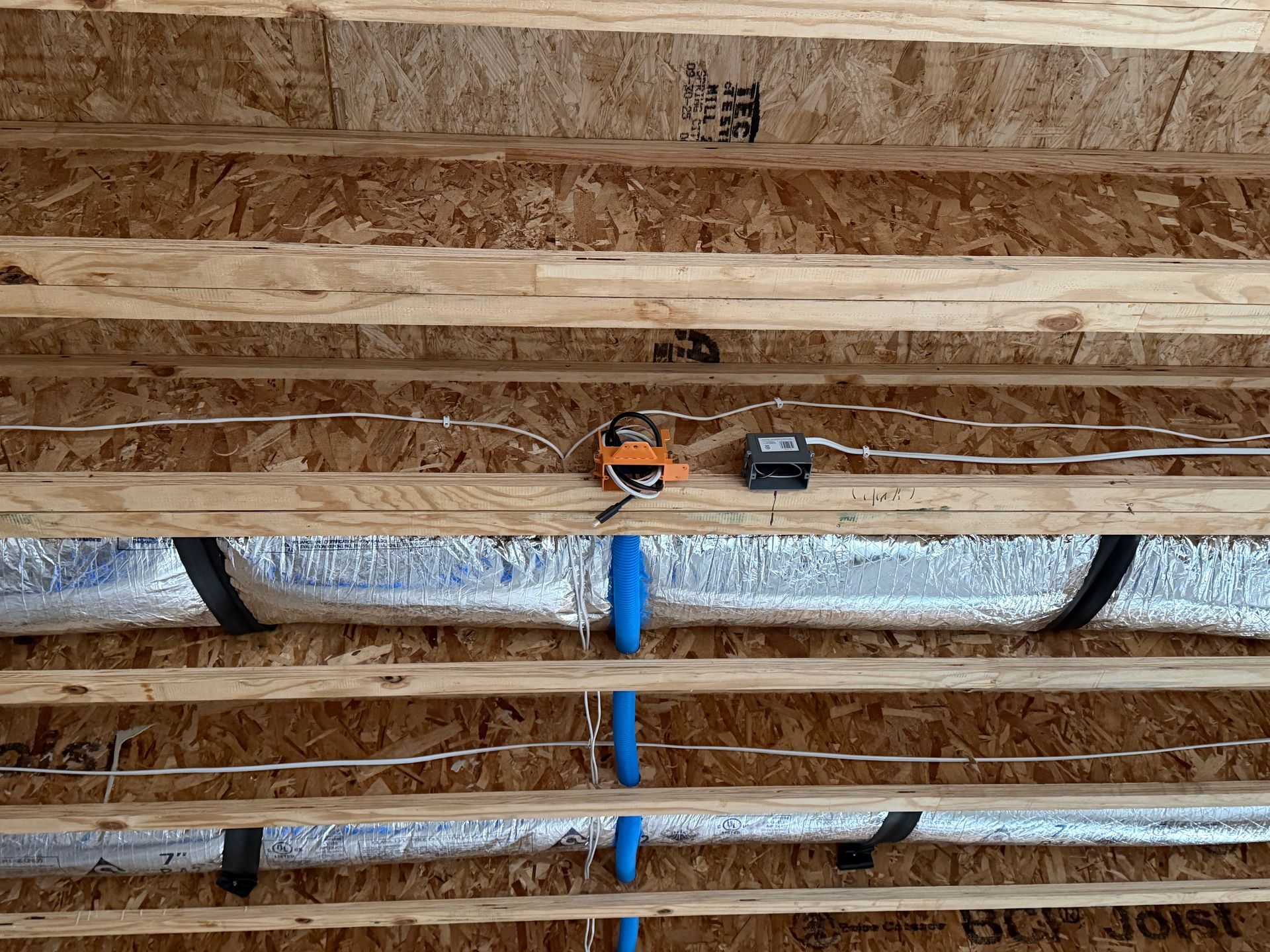 Electrical wires, a junction box, and silver insulated HVAC ducts run between parallel wooden floor joists.