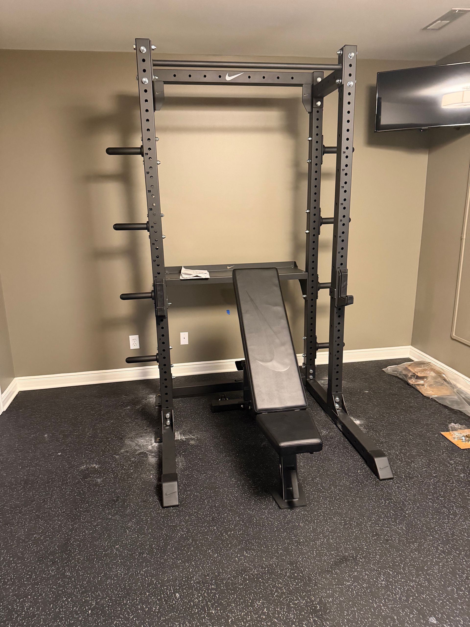 A black power rack with a weight bench inside, set on a dark, speckled gym floor against a plain wall.