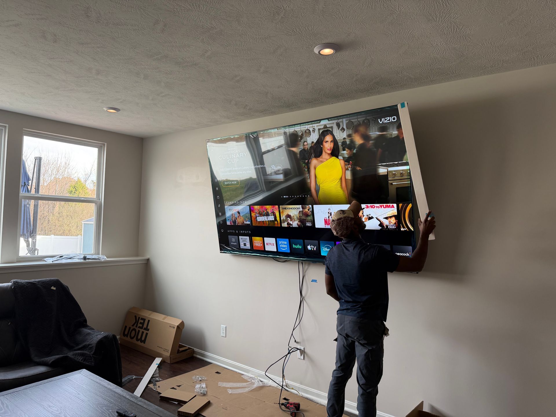 A person mounts a large television on a light-colored wall in a room with a window and cardboard packaging on the floor.