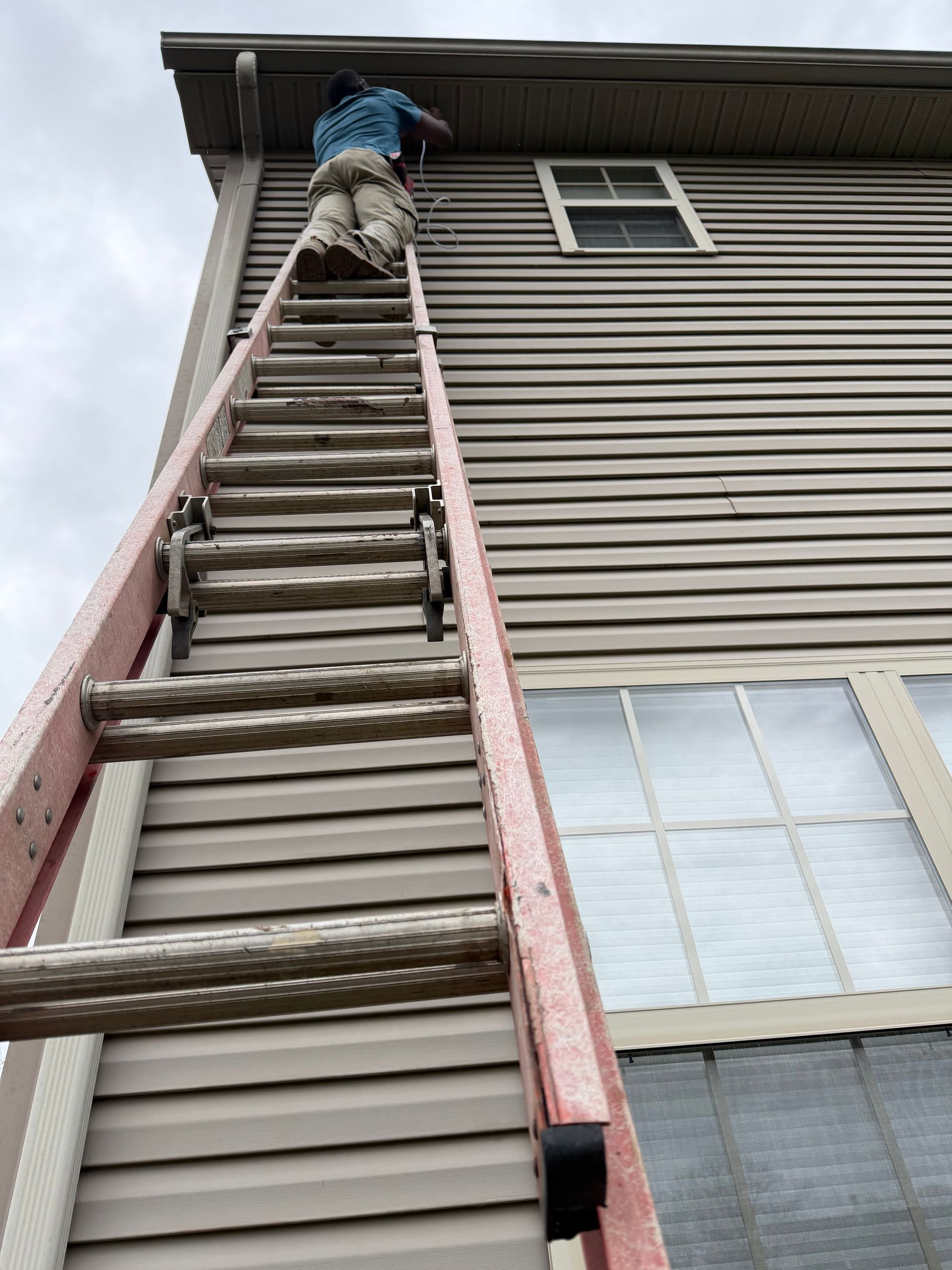 A person wearing tan pants and a blue shirt climbs an extension ladder leaned against the tan siding of a two-story house.