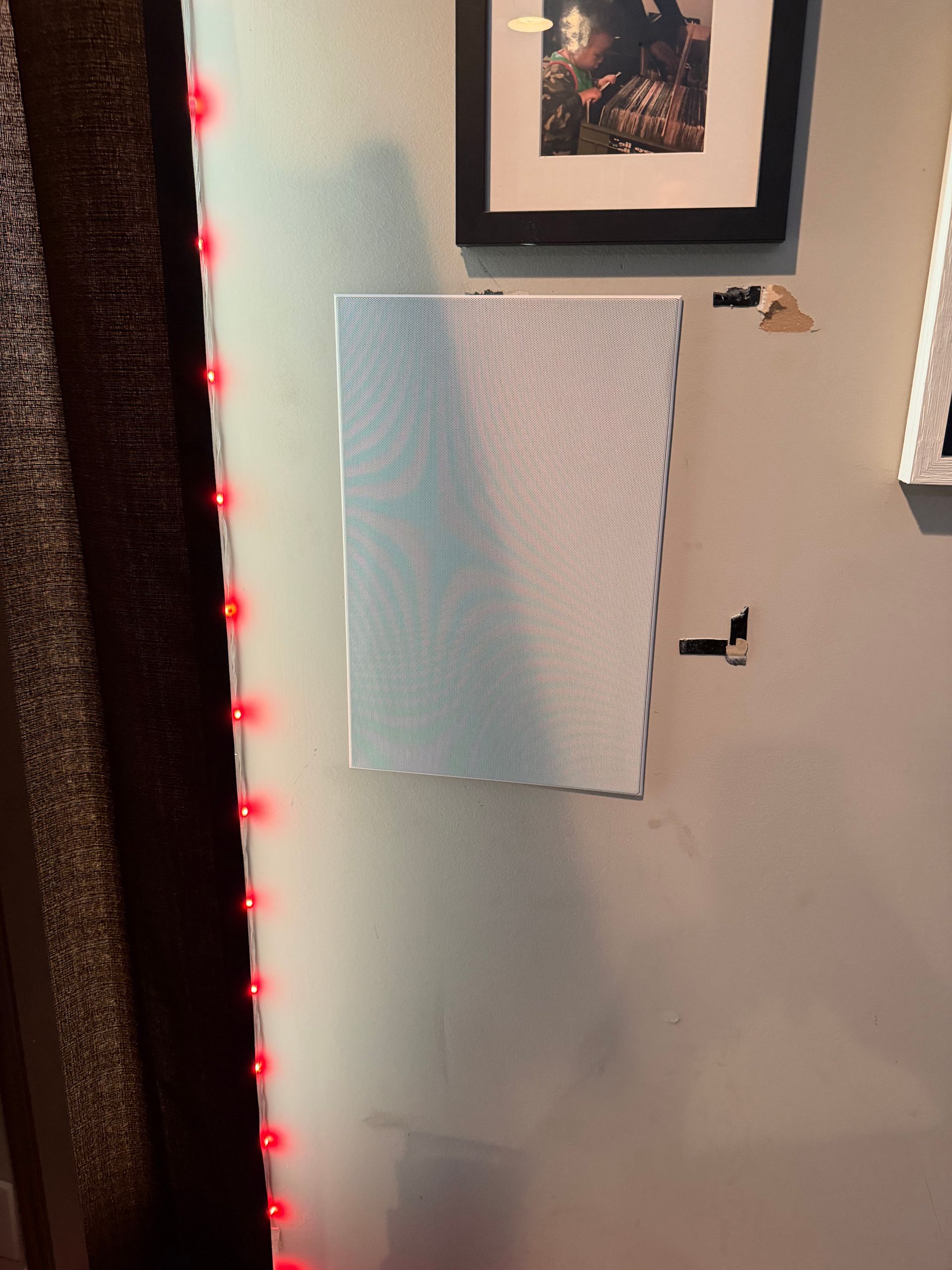 A wall-mounted speaker with a white mesh grille next to a string of red LED lights and a framed picture on a grey wall.