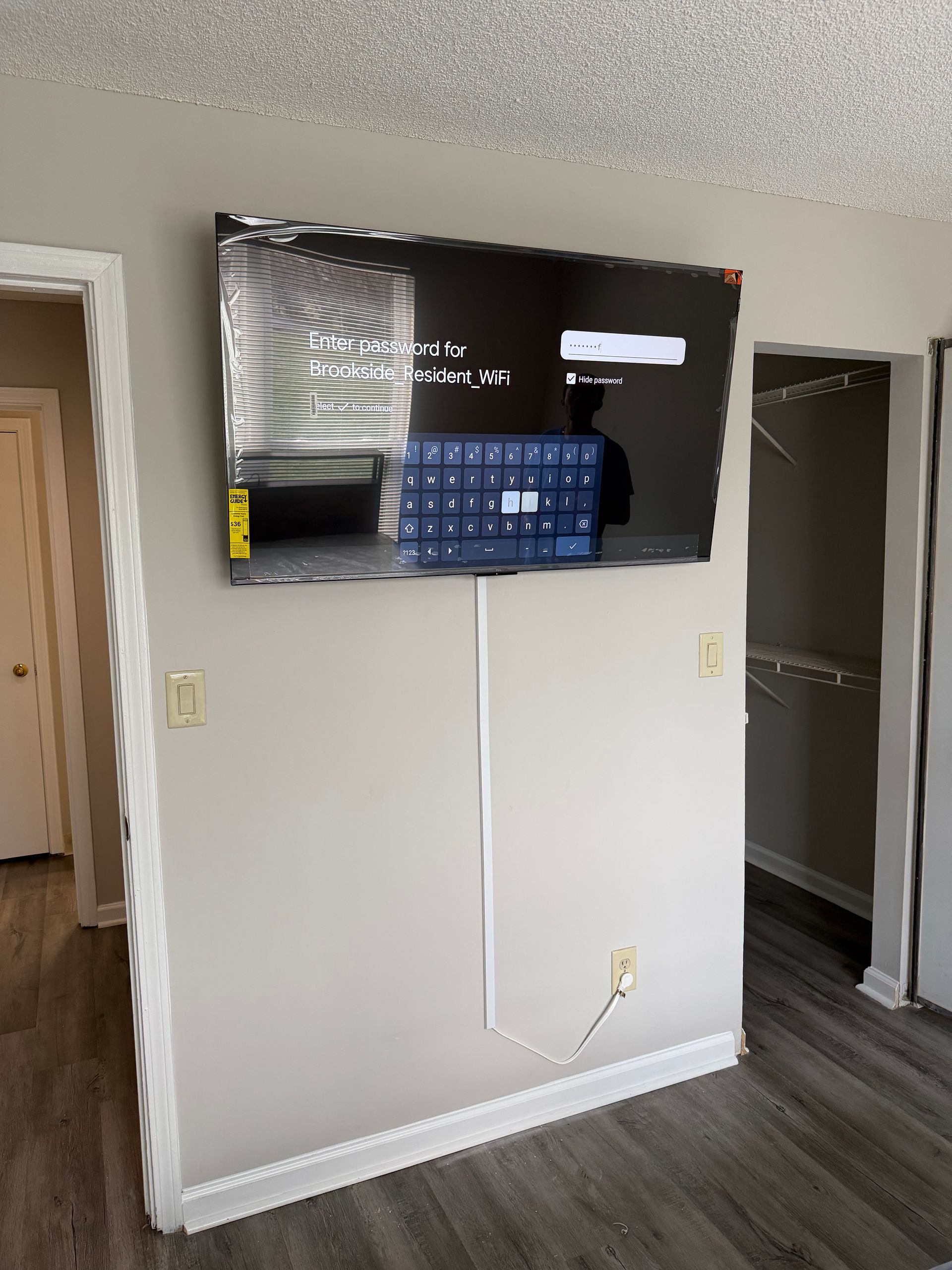 A wall-mounted TV displays a digital keyboard and setup menu in a room with light grey walls and wood-patterned flooring.