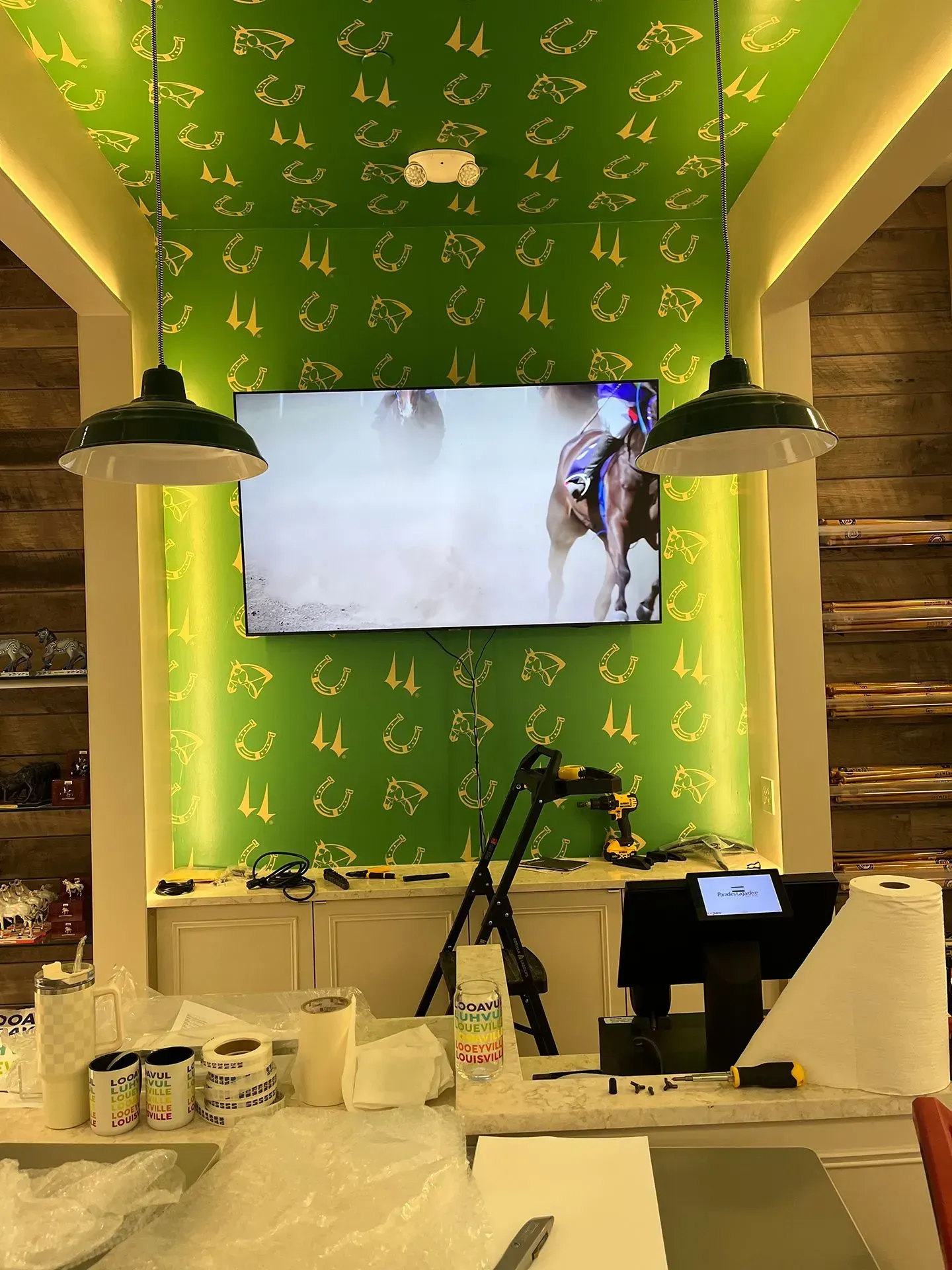 A flat-screen TV playing horse racing mounted on a vibrant green wall with gold horseshoe patterns, flanked by lamps.