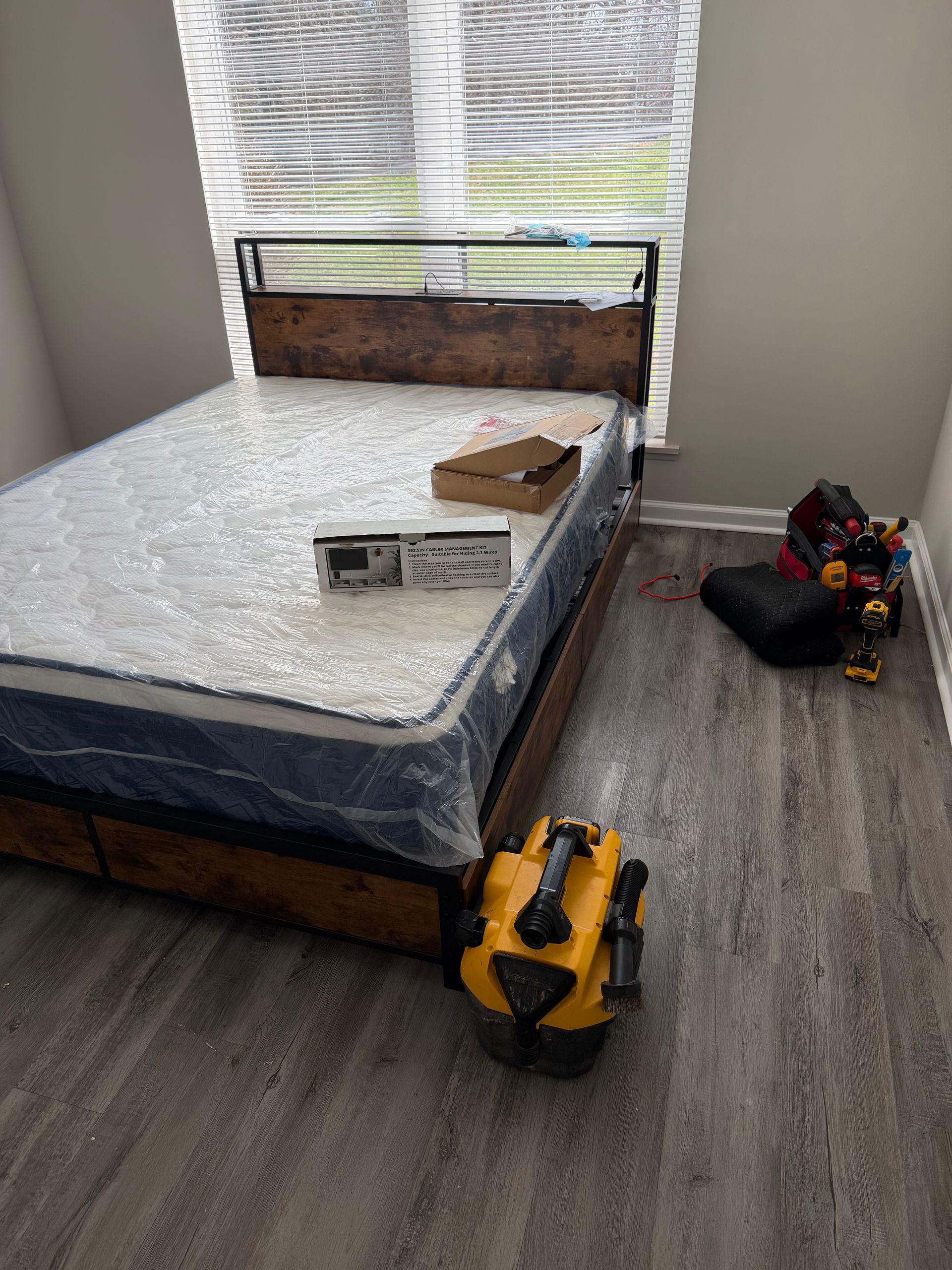 A wooden bed frame with a mattress inside a room, featuring a yellow shop vac on the floor and scattered items.