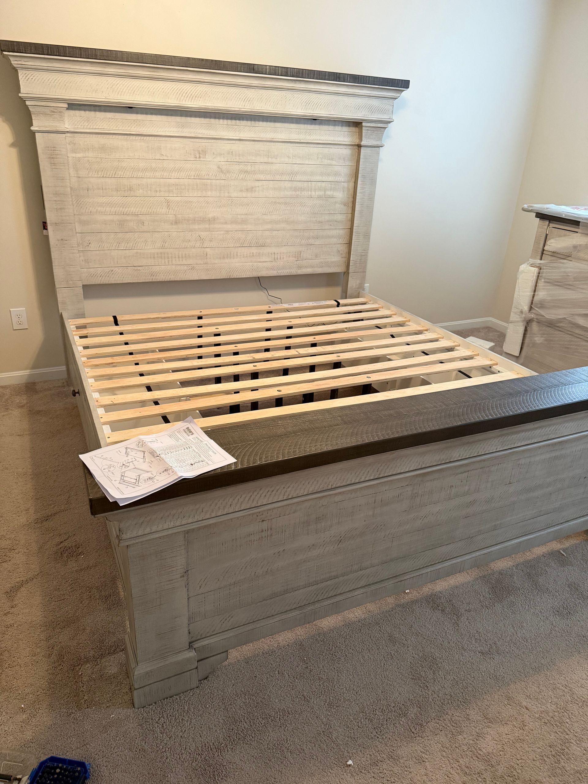 A white, rustic-style wooden bed frame with a headboard and slats sits on carpet in a room with light-colored walls.